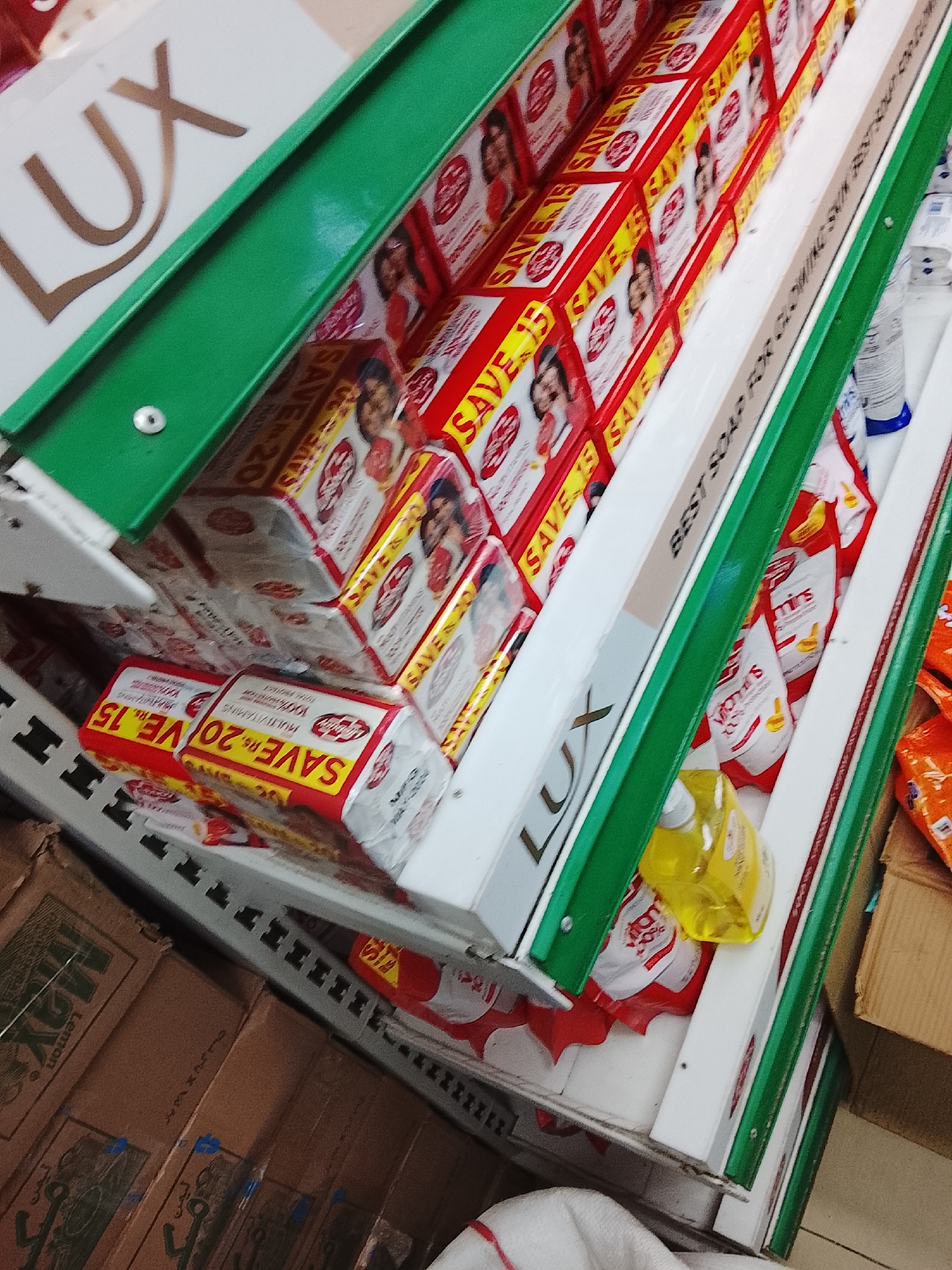 The image shows a shelf in a shop in Gulgasht Colony, Pakistan. The shelves are stocked with Lux soap, in boxes labeled "Save 15" and "Save 20". There are also bottles of what appears to be dishwashing liquid and various other products. Below the shelf is a cardboard box labeled "Max Lemon".