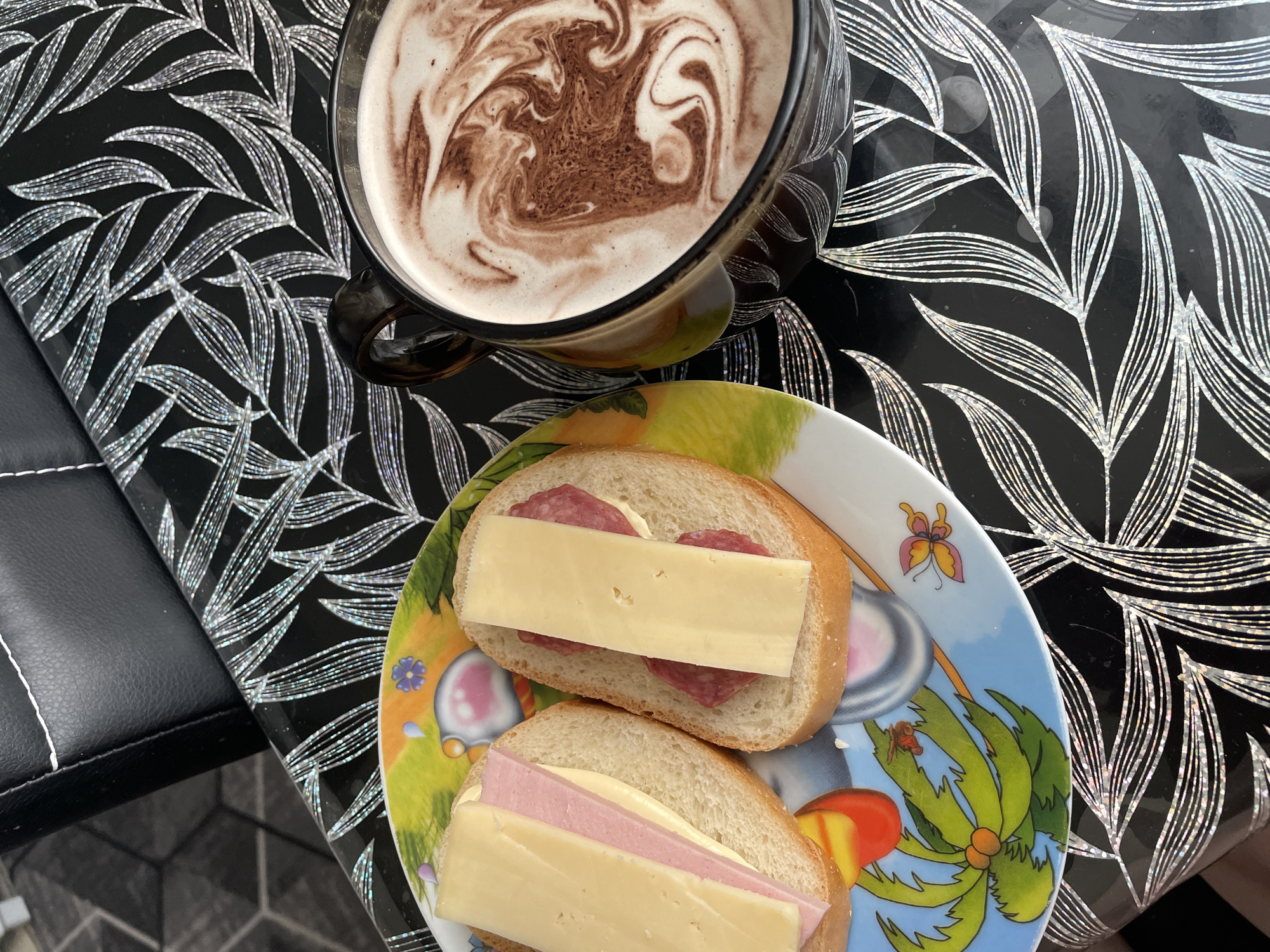 A still life is presented, featuring a mug of hot chocolate with swirled brown cocoa atop a frothy white surface, and a plate holding two open-faced sandwiches. The sandwiches appear to be on white bread with slices of processed cheese and different types of meat; one has salami and the other has ham. The mug and plate rest on a glossy black surface adorned with a leaf pattern in silver or white. The edge of what appears to be a black chair with white stitching is visible on the left. The exact location in the city of Chaikovsky, Russia cannot be determined from this image alone.