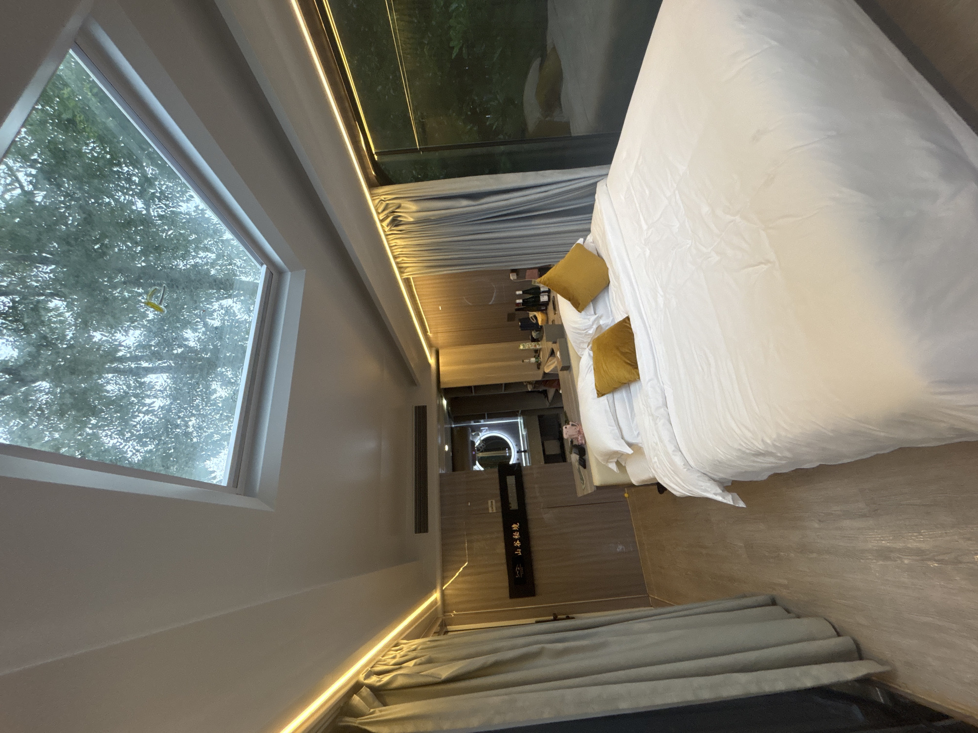 This is a wide-angle, indoor, eye-level shot of a bedroom. The room appears to be a hotel room, possibly in Guangzhou City, China, given the presence of Chinese characters on a wall plaque. The dominant feature is a large, white bed with two mustard-colored pillows in the foreground. The bed is covered with white linens and is neatly made.
Above the bed, a large skylight allows natural light to filter in, revealing the silhouettes of trees outside. The ceiling is flat and white, with warm, yellow LED strip lighting running along the edges of the walls.
To the left of the bed, a grey curtain is drawn, obscuring a doorway or window. Further into the room, a wall with wood-grain paneling is visible. On this wall, a black plaque with gold Chinese characters, possibly a hotel name or slogan, is mounted. Next to the plaque is a circular mirror, and behind it, a glimpse of a sink and countertop suggests a bathroom area.
To the right of the bed, a large window or glass door offers a view of lush greenery outside, indicating a natural or garden setting. The floor is a light-colored wood laminate. On a surface next to the bed, several bottles and cups are visible. The overall impression is a modern, comfortable, and nature-inspired accommodation.