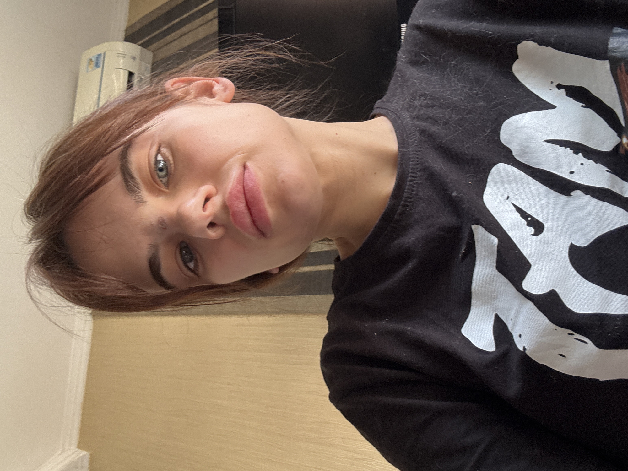 A young woman is pictured indoors. She has fair skin, light brown hair, and blue eyes. She wears a black t-shirt with a large white graphic. The background includes a beige wall and a white appliance. There is no evidence indicating that the location is in Rostov-on-Don, Russia.