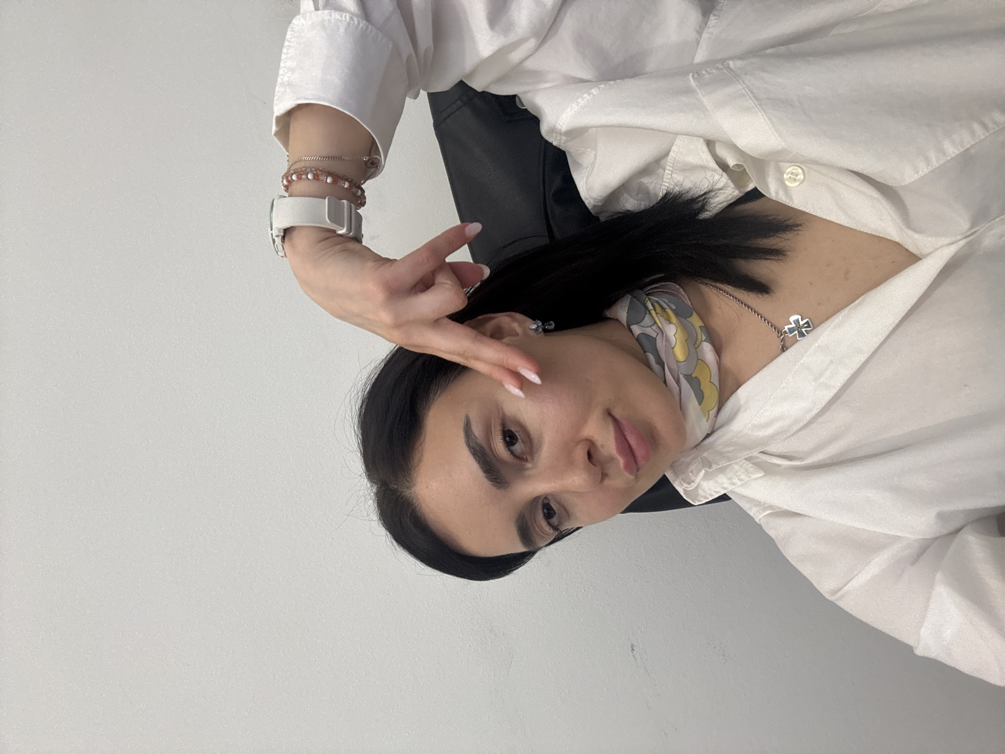 A medium, rotated, eye-level shot of a woman with long black hair and fair skin, wearing a white button-up shirt, seated in a black leather chair against a light gray wall. She is facing the camera and making a "V" sign with her fingers near her left temple. She is also wearing a white smartwatch, multiple bracelets, a floral neck scarf, and a silver cross necklace. The location is likely indoors, but not identifiable as Tbilisi, Georgia.