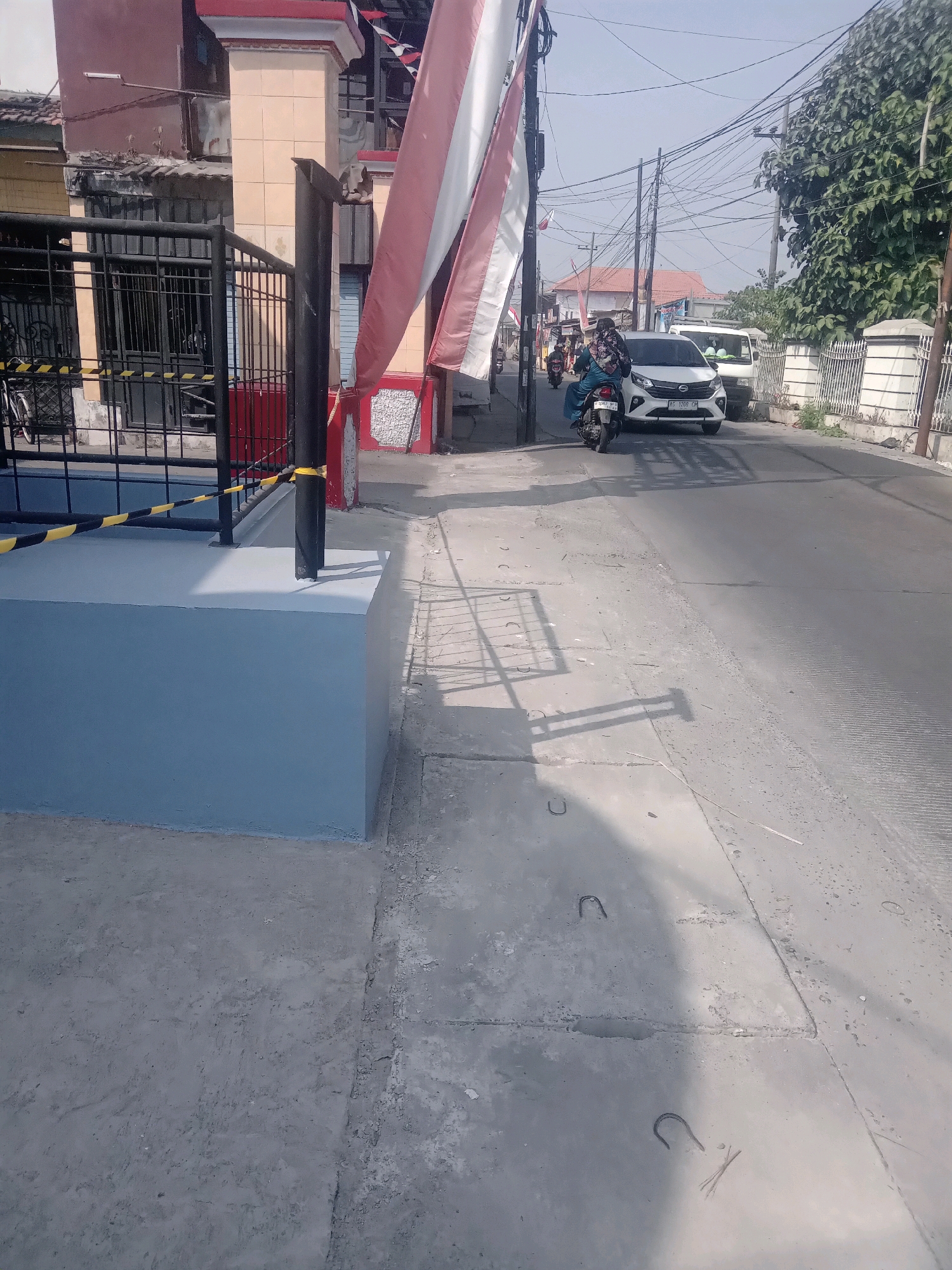 The image depicts a street scene in Wage, Indonesia, likely on a bright, sunny day given the strong shadows. The foreground features a light blue, rectangular concrete structure with a black metal pole rising from its center, adorned with yellow and black striped tape. Adjacent to this structure is a black metal fence with vertical bars and a gate. To the left, a bicycle is partially visible leaning against the fence.
The street itself is paved with asphalt and concrete. A white car is parked on the right side of the road, and a motorcycle carrying two people is moving away from the camera on the left lane. The individuals on the motorcycle appear to be wearing modest clothing, with one person wearing a headscarf. Beyond the street, a white van is visible.
The background includes buildings with red and cream colored facades, some appearing aged. Overhead, a complex network of electrical wires crisscrosses the sky, supported by wooden and concrete poles. Lush green trees line the right side of the street, and a white decorative fence runs along the property line. Indonesian flags, red and white, are displayed on poles, indicating a national celebration or presence. The overall impression is a typical residential or commercial street in a moderately developed Indonesian town.