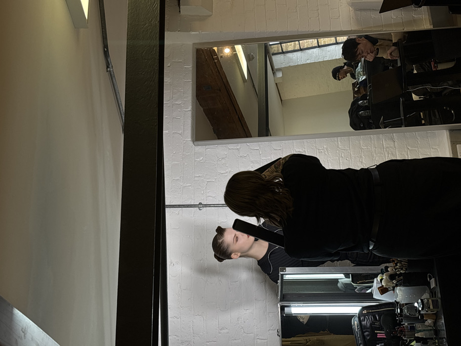 The image depicts an indoor studio setting in a building consistent with industrial conversions found in urban areas of London, United Kingdom. The immediate background features a white-painted brick wall with visible dark metallic electrical conduit running vertically. A substantial dark, textured metal beam spans horizontally across the upper portion of the frame.
To the left, a vanity or makeup station is present, comprising a dark case opened to reveal two vertical fluorescent lights and numerous makeup brushes and containers. Standing against the brick wall, a young individual, serving as a model, is positioned with light hair styled into two prominent upward buns. They wear a dark, ribbed garment and exhibit a neutral expression with eyes partially closed, their face turned slightly to the left.
In the center foreground, viewed from behind, a person with medium-length dark hair and visible arm tattoos is operating a dark photographic or video camera, holding it to their eye level. They are dressed in dark clothing.
To the right, a large, rectangular mirror with a thin white frame reflects another segment of the room. This reflection reveals two additional individuals: one partially obscured on the far right, and another seated at a table, leaning forward with a hand supporting their chin while observing a laptop. The reflection also shows an exposed wooden ceiling beam and a window or interior architectural element.