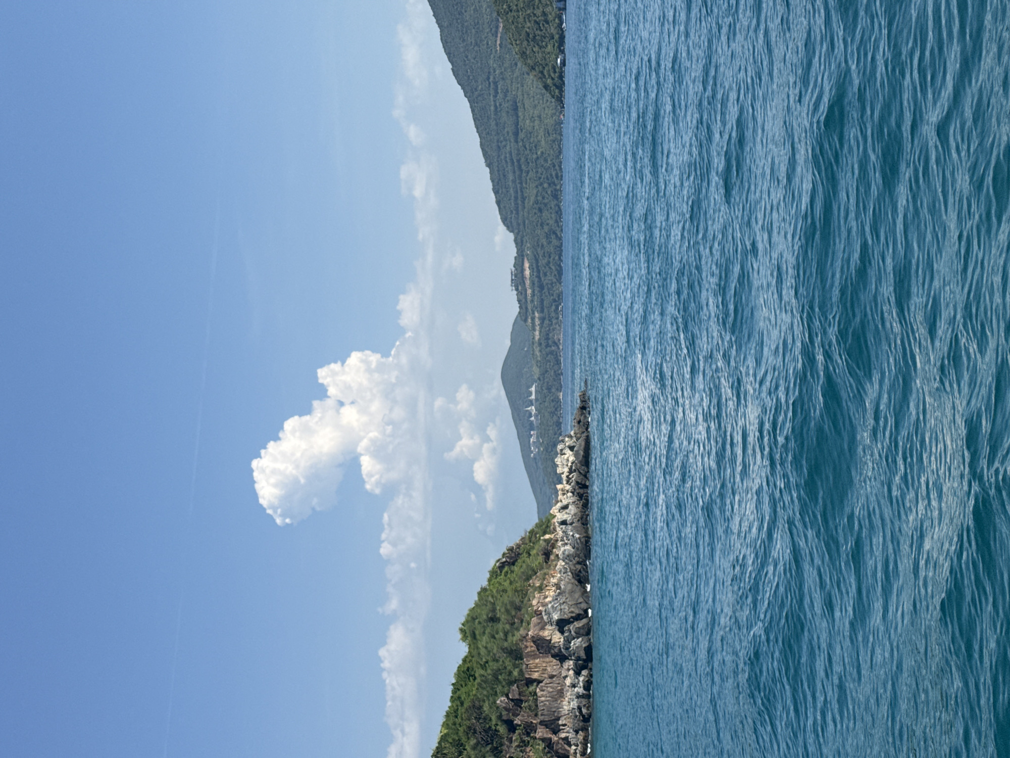 The image depicts a scenic view of the sea and coastline. In the foreground, the blue sea is slightly choppy with visible waves. The mid-ground features a rocky island on the left, covered with dense green vegetation and dark rocks. Further to the right, the coastline is characterized by green, sloping hills that recede into the distance. The sky above is a clear blue with a few white clouds, including a prominent, unusually shaped cloud in the upper center. In the far distance, on the slopes of the hills, there appear to be some structures, possibly buildings or resorts, with white roofs. The overall scene suggests a tropical or subtropical coastal environment. This appears to be a view from a boat or a coastal vantage point looking out towards the sea and islands. The location is identified as being in Nha Trang Ward, Vietnam, which is known for its beautiful beaches and islands.