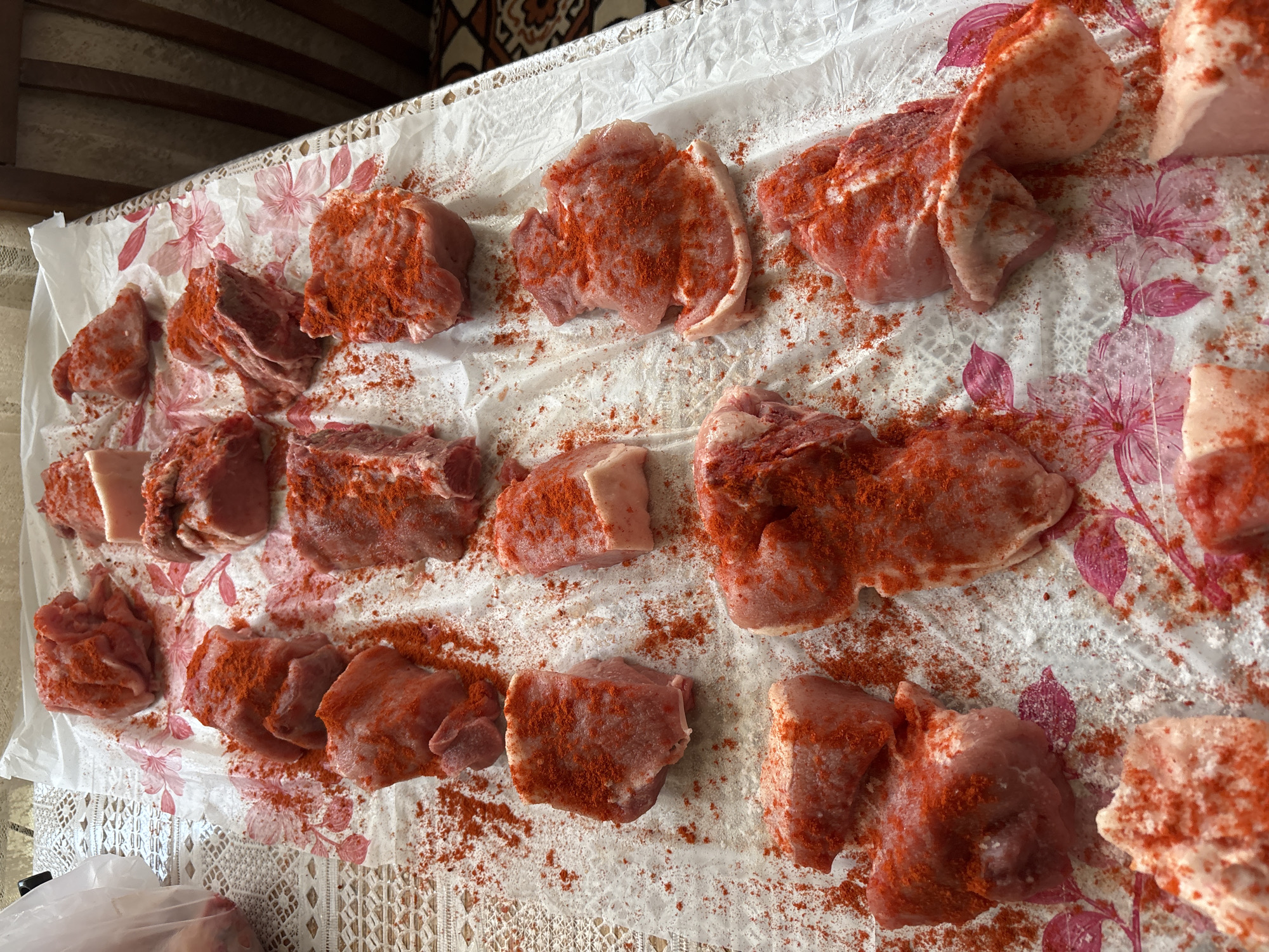 The image captures an overhead view of numerous pieces of raw meat, likely pork, meticulously laid out on a patterned surface and generously seasoned with a bright red spice, possibly paprika or chili powder. The meat cuts vary, with some appearing as lean chops, others as chunks with visible fat, and some showing bone.
The meat is arranged on a crinkled white paper or thin plastic sheet adorned with a repeating pattern of pink and red flowers. This sheet, in turn, covers a more elaborate, white, lace-patterned tablecloth. A substantial amount of the red spice is scattered not only on the meat but also across the white paper, suggesting it was recently applied during the seasoning process.
The scene depicts an activity of food preparation, likely for a meal, or possibly for curing or preservation. The background reveals a dark, possibly wooden, ceiling structure or beam, indicating an indoor domestic setting. No people are visible, nor is any text present in the image. The overall impression is one of careful culinary preparation in a home in Vagharshapat, Armenia.