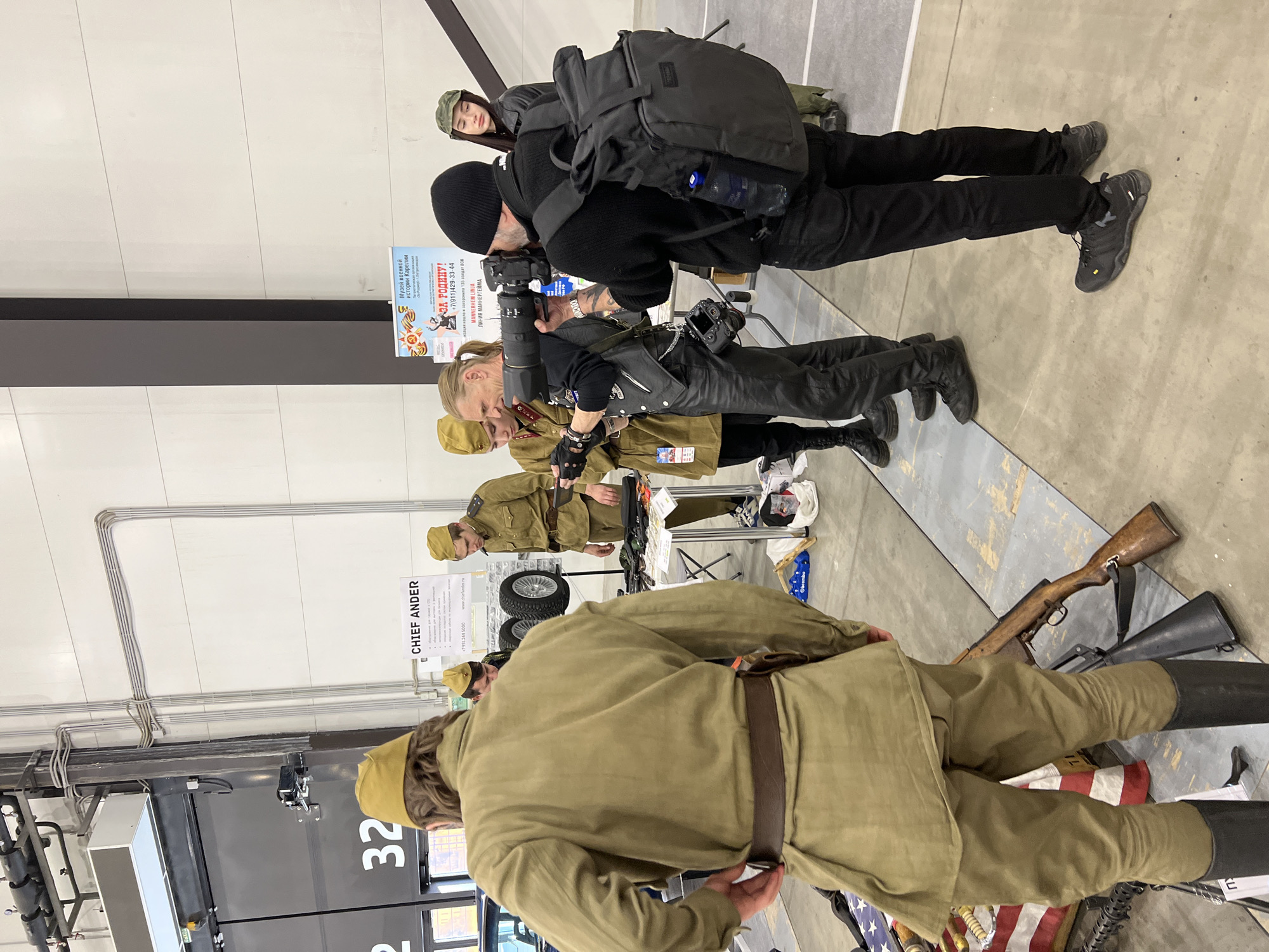 Here is a description of the image:
The image shows a scene indoors, seemingly at an event or convention, possibly in Shushary, Russia. There are several people present, some dressed in what appear to be military uniforms from an earlier era, possibly Soviet uniforms, and others dressed in modern clothing. The uniformed individuals are standing around a table with what looks like equipment, possibly related to military history. A photographer is taking pictures of them. The environment includes event signage, indicating possible museum or historical affiliations, such as "Музей военной истории Карелии" and "MANNERHEM LINJA". A sign is also visible that contains the name "CHIEF ANDER". A firearm is visible. The overall setting appears to be an indoor event focused on military history or reenactment.