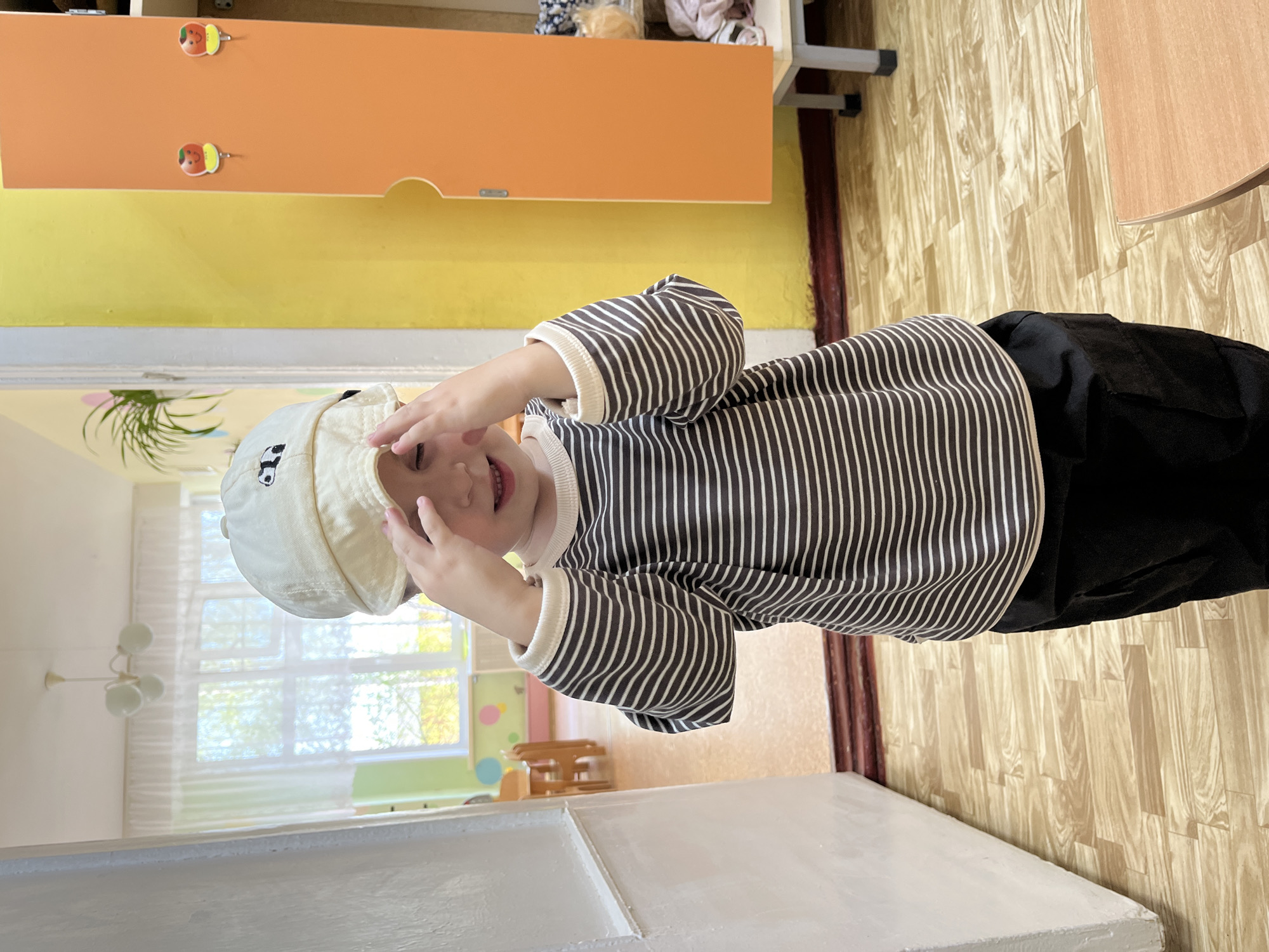 A child in a striped shirt and cap stands indoors. The child holds their hands near their face. The room has orange and yellow walls and is located in Chaikovsky, Russia. The room also features a wooden floor.