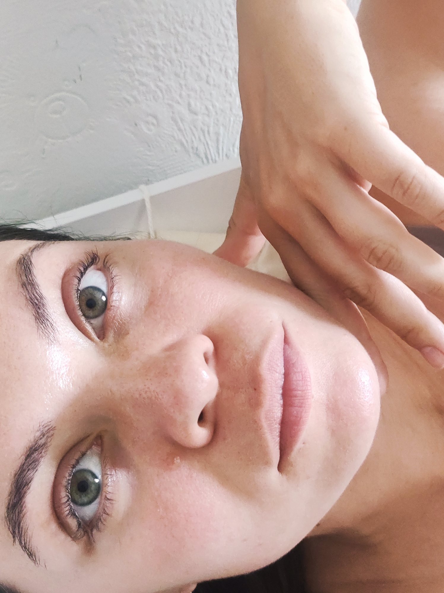 This media file presents a close-up portrait of a woman, captured from a slightly upward angle. Her face occupies the left side of the frame, with a hand, likely her own, gently resting on her right shoulder and neck on the right side of the image.
The woman has fair skin, which appears dewy or moisturized, and visible freckles across her nose and cheeks. She possesses striking green or hazel eyes that are wide open and directed towards the viewer. Her eyebrows are dark and well-defined, complementing her dark hair, which is visible framing her face. Her lips are a natural pink hue, slightly parted.
The background primarily consists of a white, textured ceiling featuring subtle embossed patterns, including faint circular designs, visible in the upper left. A thin white line, possibly indicating a shelf or a bed frame, runs horizontally across the upper mid-section. The lighting is soft and ambient, suggesting an indoor setting. The woman's expression is calm and neutral. There is no visible text in the image. This scene is set in an indoor location in Chaikovsky, Russia.