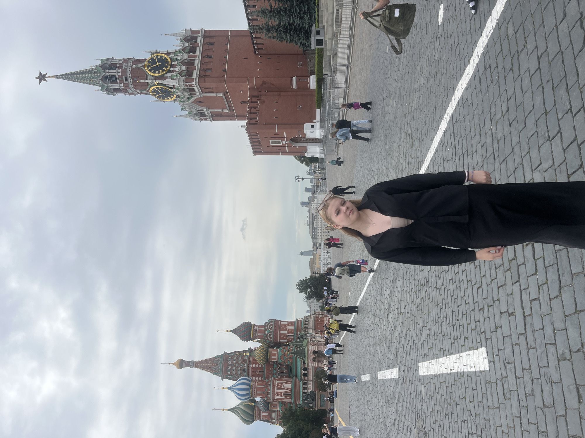 This image captures a street scene in Moscow, Russia, specifically Red Square. In the foreground, a young woman with blonde hair is posing for the camera, wearing a black button-up shirt and a black skirt. She has a light-colored top underneath her shirt. Behind her, the iconic St. Basil's Cathedral, with its colorful onion domes, is visible on the left. On the right, a tall brick tower, likely the Spasskaya Tower of the Moscow Kremlin, stands prominently with a star on top and large clock faces. The ground is paved with cobblestones, and white lines mark a path or road. Several other people are scattered throughout the square, some walking and some standing. The sky is overcast with clouds. A large green duffel bag is being carried by someone partially visible on the right side of the image.