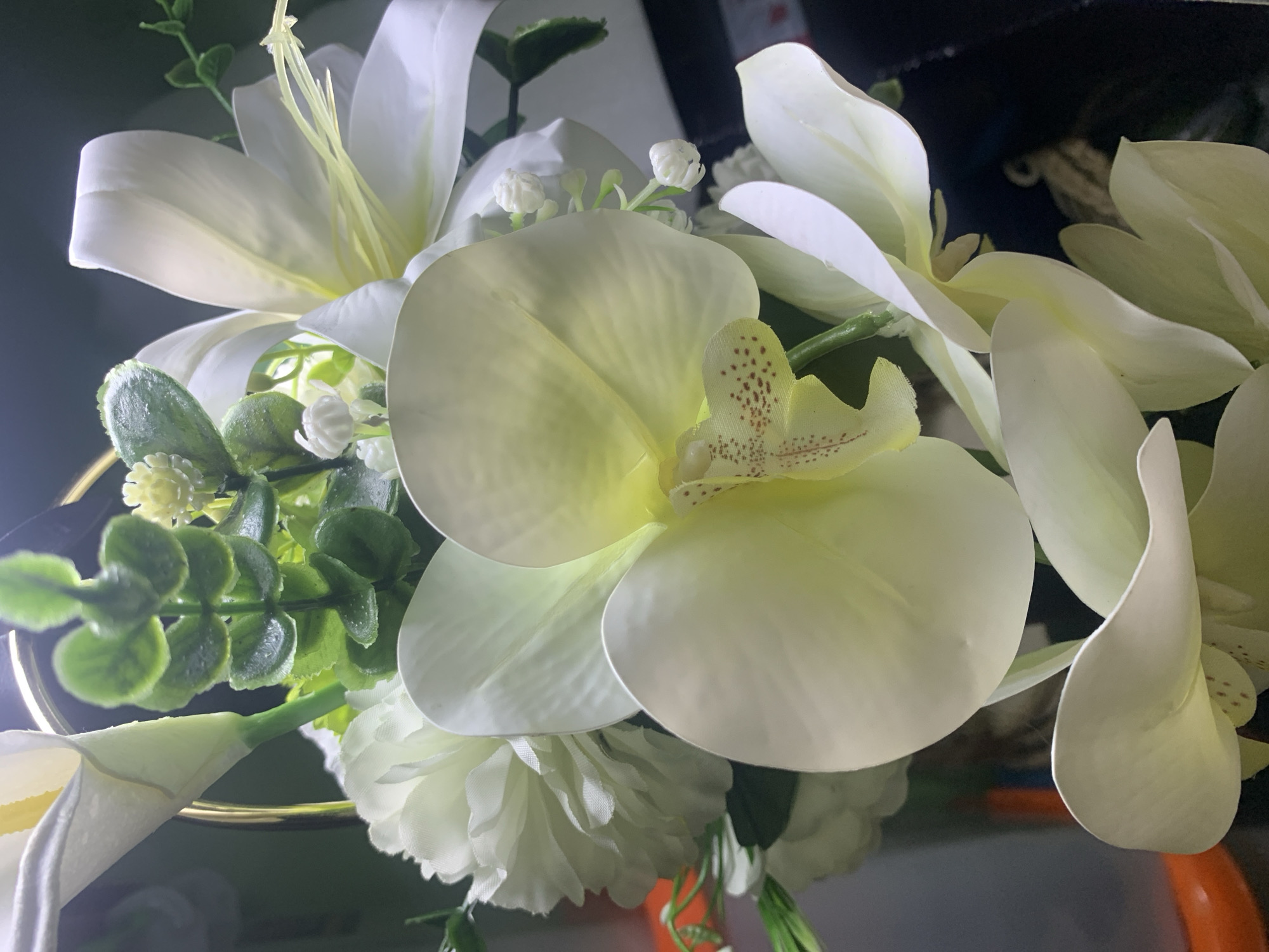 This is a close-up, indoor photograph of an artificial floral arrangement. The primary subjects are several large, pale yellow orchid blooms with speckled centers, interspersed with white lily-like flowers and clusters of small green leaves. The lighting is dramatic, with bright highlights on the petals and dark shadows in the background, suggesting artificial lighting rather than natural sunlight. The arrangement appears to be placed in a vase or container, with a hint of a gold rim visible at the bottom left. There are no people, text, or specific location cues other than the given location context. The scene evokes a sense of decorative elegance, with the artificial nature of the flowers being evident upon closer inspection of their texture and uniformity. The focus is entirely on the detailed textures and forms of the flowers and foliage.
