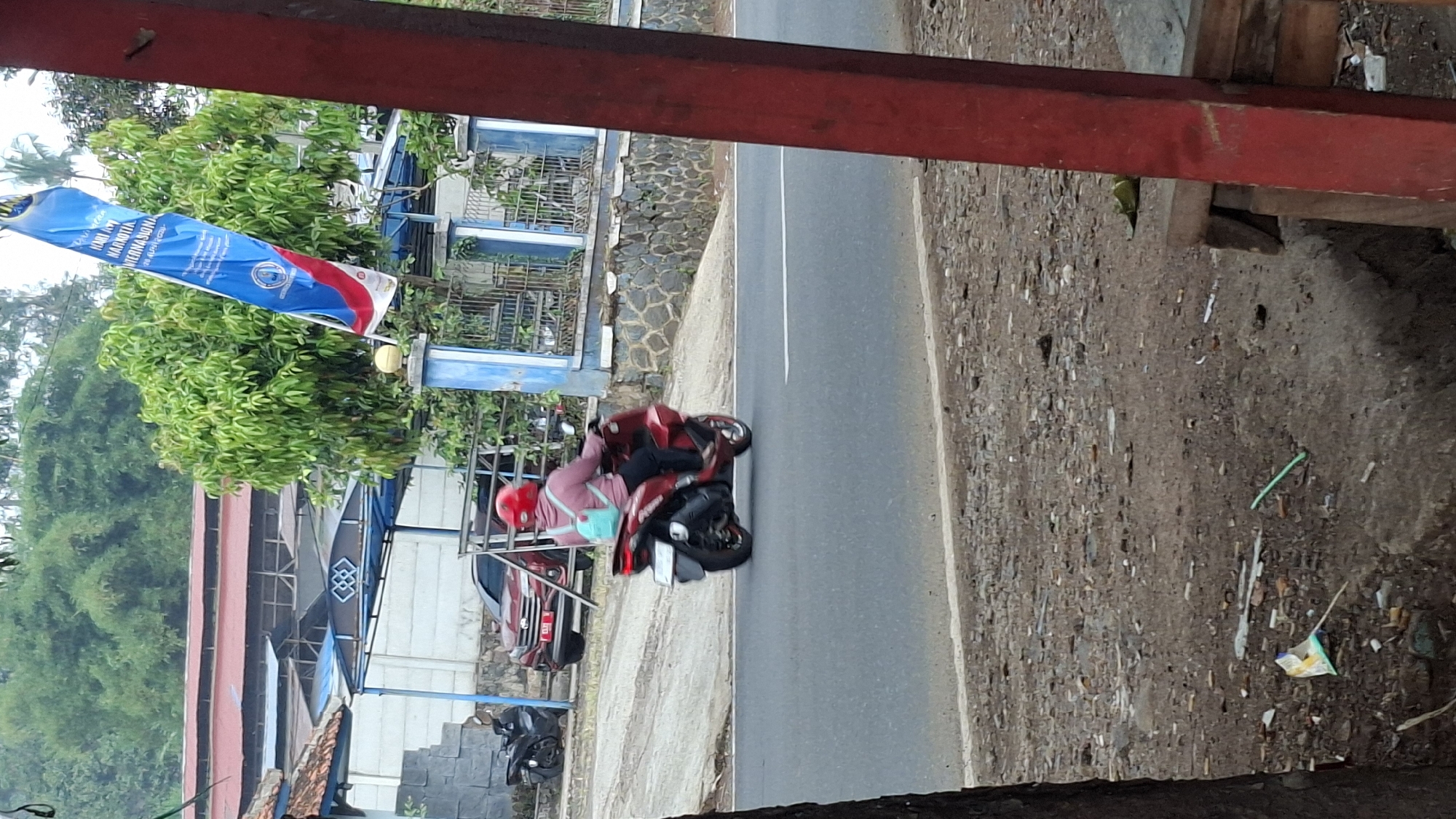 The image depicts a street scene in Sirnagalih, Indonesia. A person wearing a red helmet and a pink jacket is riding a red motorcycle on a paved road. The person is carrying a light blue backpack. To the left of the motorcycle, a black scooter is parked. In the background, there is a red car parked behind a fence. Further back, there are buildings with red roofs and blue awnings. A tall, blue and white banner with text in Indonesian hangs from a pole. The ground on the side of the road is covered in dirt and debris, including discarded cigarette butts. A red metal post is visible in the foreground on the right.