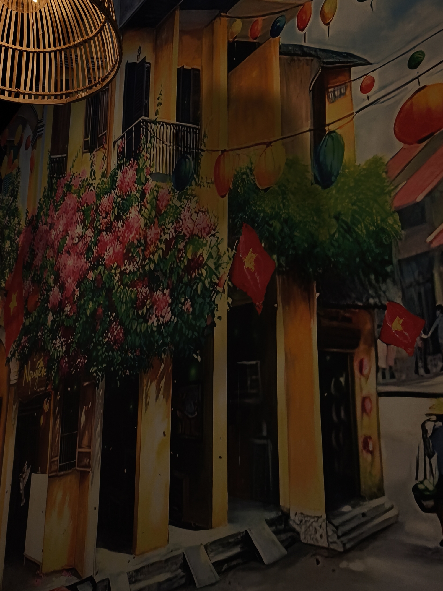 The image depicts a mural painted on a wall, showcasing a vibrant street scene. The mural features buildings with yellow walls and dark shutters, adorned with a profusion of pink flowers and lush green foliage. Several red Vietnamese flags with yellow stars are prominently displayed. Overhead, a string of colorful lanterns adds to the festive atmosphere. In the foreground, a bamboo light fixture hangs, partially obscuring the mural. The overall style is reminiscent of a Southeast Asian setting. There is no indication within the image or its context that this mural is located in the city of Izhevsk, Russia. The image captures a static, painted scene and does not contain any observable people, actions, or interactions.
