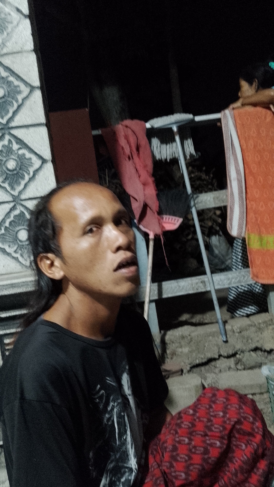 The image captures a close-up of a bald man with dark hair pulled back, wearing a black t-shirt. He is looking upwards with his mouth slightly open. In the background, a clothesline is visible with various items of clothing hanging, including a red cloth and an orange towel with white stripes. A broom with a red handle leans against the clothesline. The setting appears to be outdoors at night, with a wall decorated with a patterned relief on the left side of the frame. The ground is rough and uneven. The man's t-shirt has a graphic print on it. The overall scene suggests a humble domestic environment, possibly in a rural area like Seputih, Indonesia.