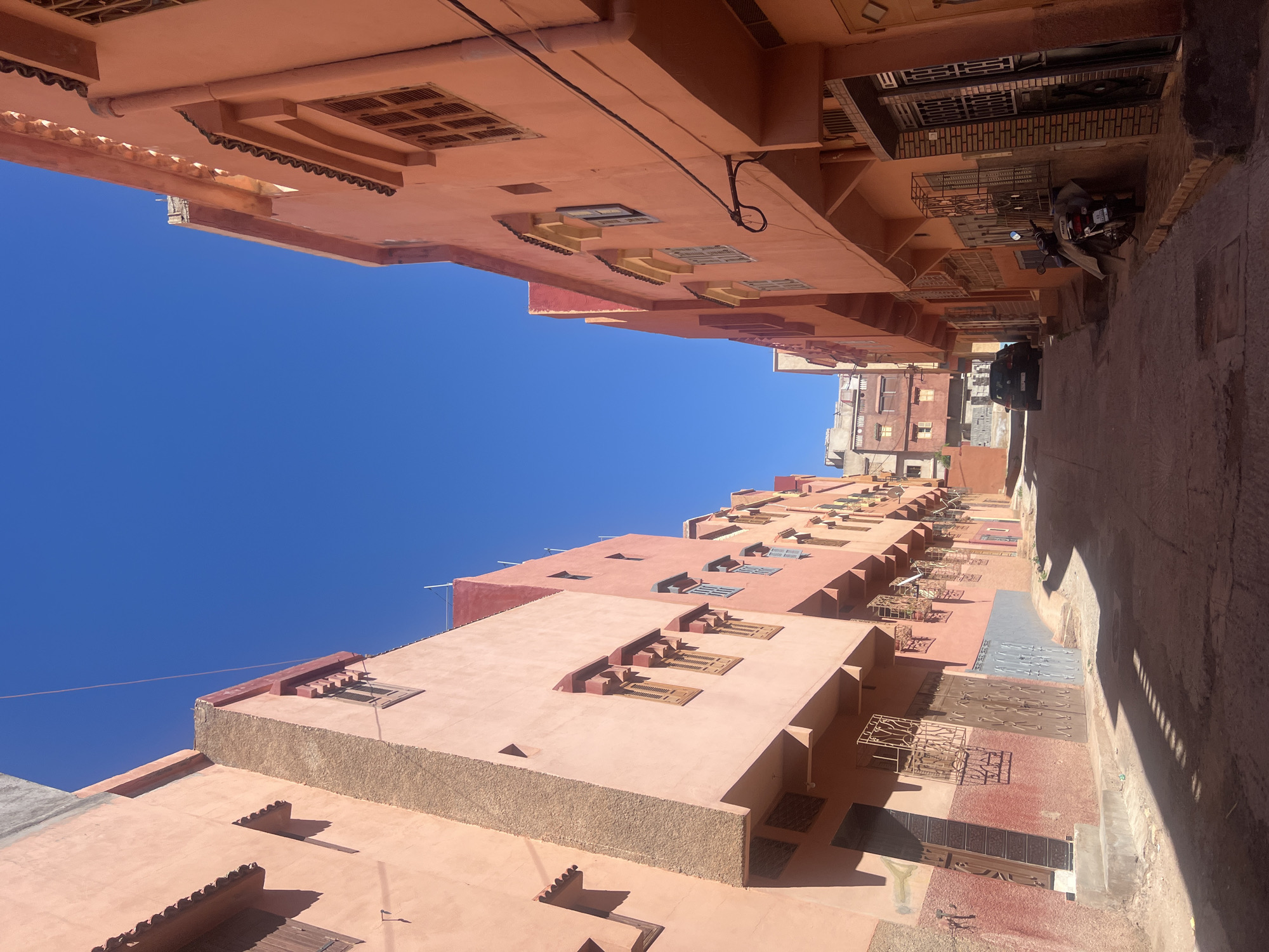 An eye-level shot of a street in Ouaouizert, Morocco, looking upward towards a cloudless blue sky. The buildings are predominately constructed of terra cotta colored stone with horizontal windows. The street below is narrow with a vehicle and motorcycle traveling away. The street is heavily shadowed on the right and sunny on the left.