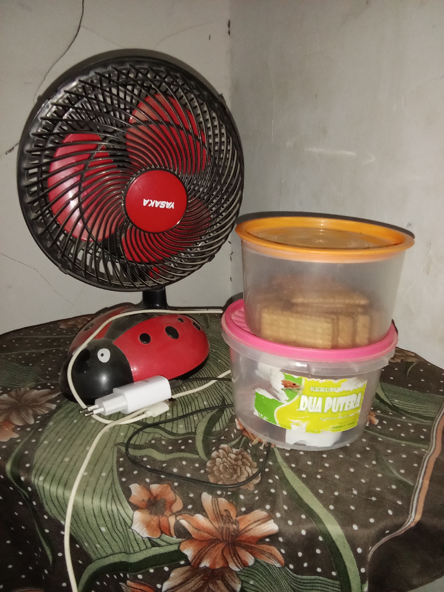 The image displays a tabletop scene with various objects. On the left, a red and black oscillating fan with the brand name "YASAKA" visible on its center is present. In front of the fan, a red ladybug-shaped object, likely a novelty item or accessory, is visible with a white charging plug connected to it. To the right of the ladybug object, two clear plastic containers are stacked. The upper container has an orange lid and contains what appears to be stacked cookies or crackers. The lower container has a pink lid and a yellow label with green accents, partially obscured, with text that includes "KERUPU DUA PUTERA". The tabletop is covered with a patterned cloth featuring floral motifs in shades of green, brown, and orange. The background is a plain, light-colored wall. The lighting suggests an indoor setting, possibly at night or in a dimly lit room. The scene is located in Selowogo, Indonesia.
