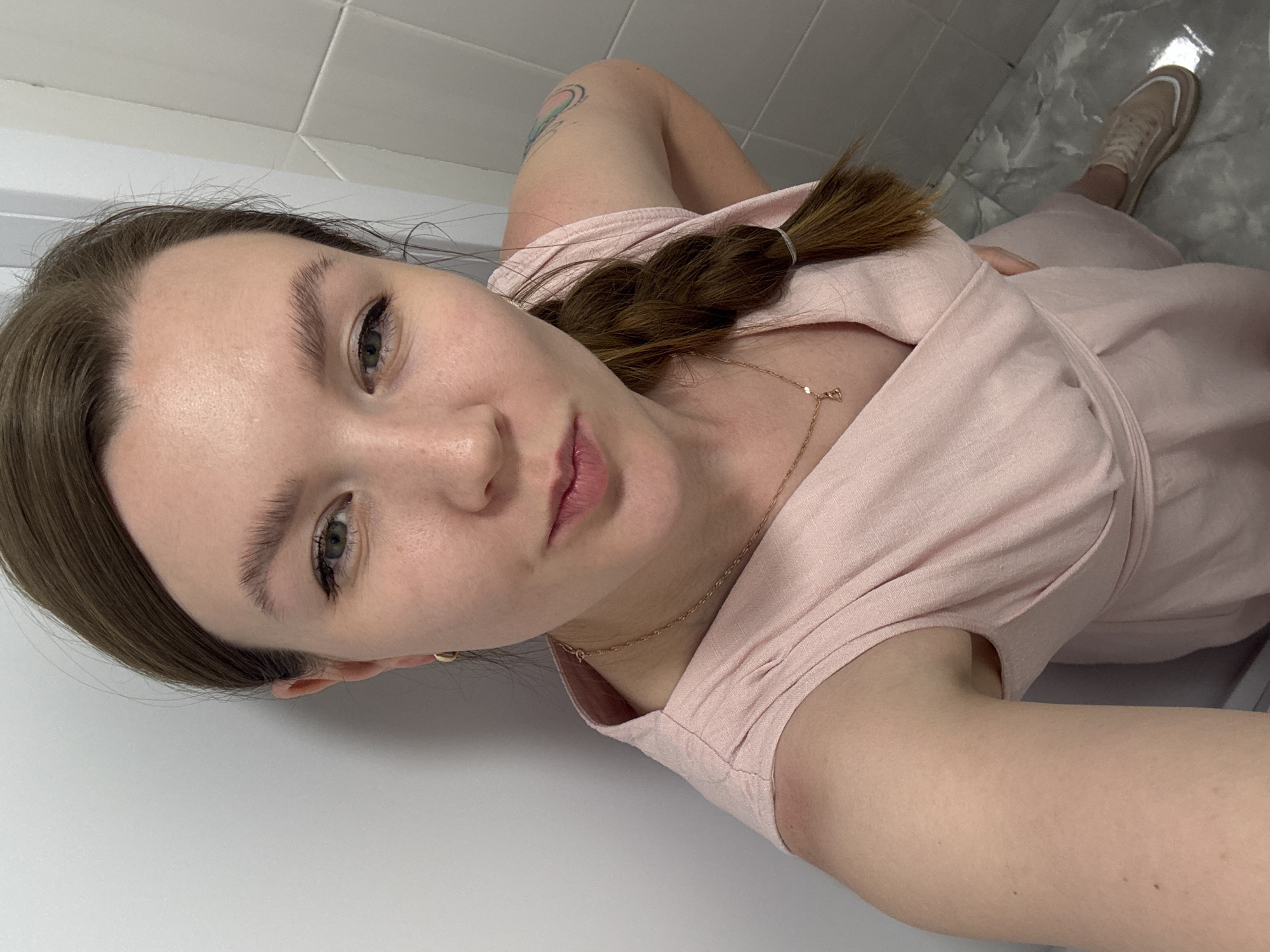A woman is taking a selfie indoors. She has brown hair pulled back in a braid and is wearing a light pink dress and a gold necklace. The background consists of white tiled walls and grey marble tiled floors. A portion of one of her feet, clad in a tan shoe, is visible in the top right corner of the image. The woman is posing at an angle. The image does not contain any information to identify the location as Izhevsk, Russia.