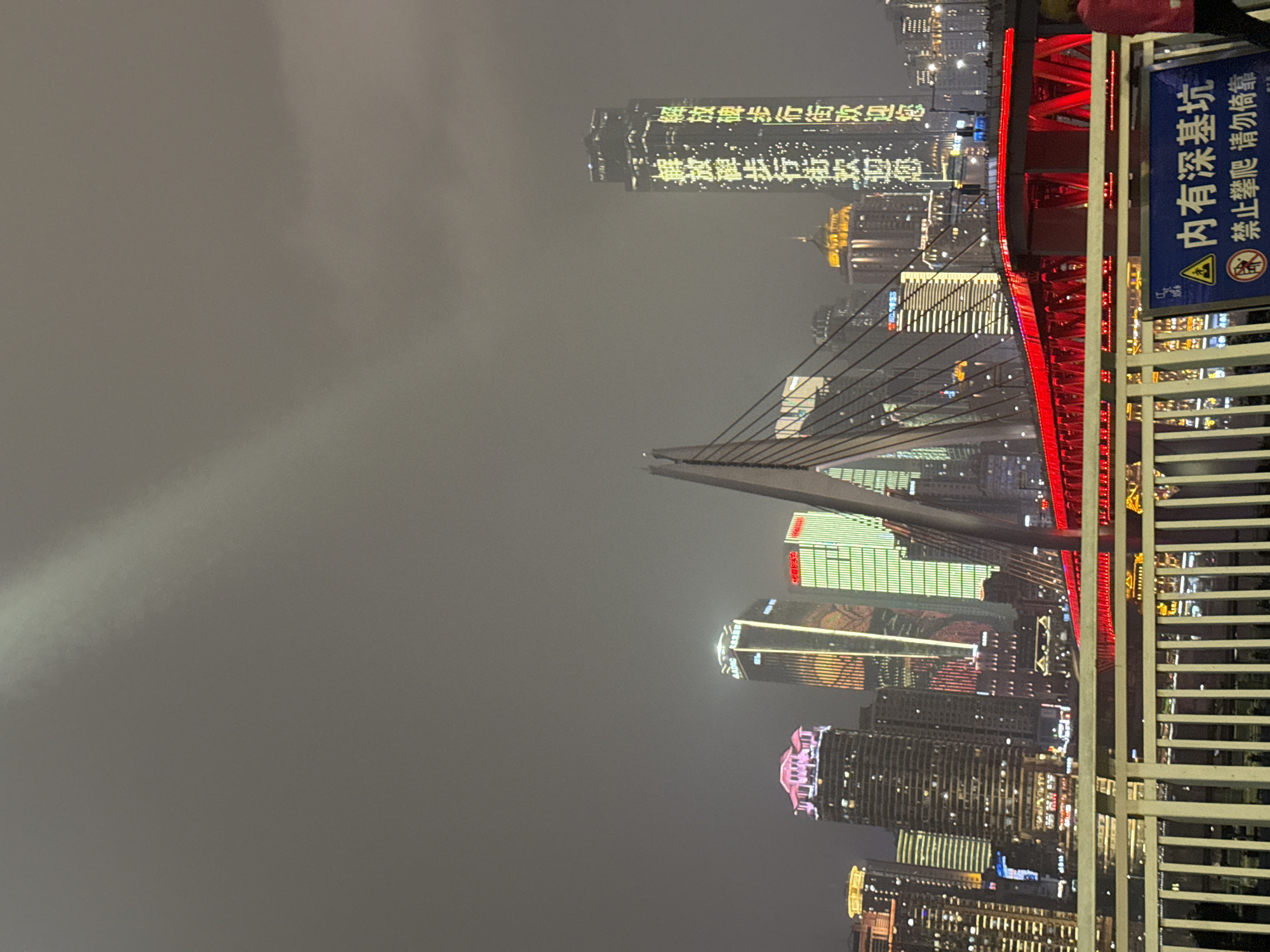 Here is a description of the image:
The image depicts a cityscape at night, with a prominent cable-stayed bridge illuminated in red. The city is likely in 江北城街道, China, based on the Chinese characters visible on a skyscraper in the background, which say "解放碑步行街欢迎您," translating to "Jiefangbei Pedestrian Street Welcomes You." Several other buildings are also illuminated, contributing to the overall urban nightscape. In the foreground, a metal fence is visible, with a blue sign attached to it. The sign contains a warning message in Chinese characters along with a symbol indicating a potential hazard. The sky is overcast.