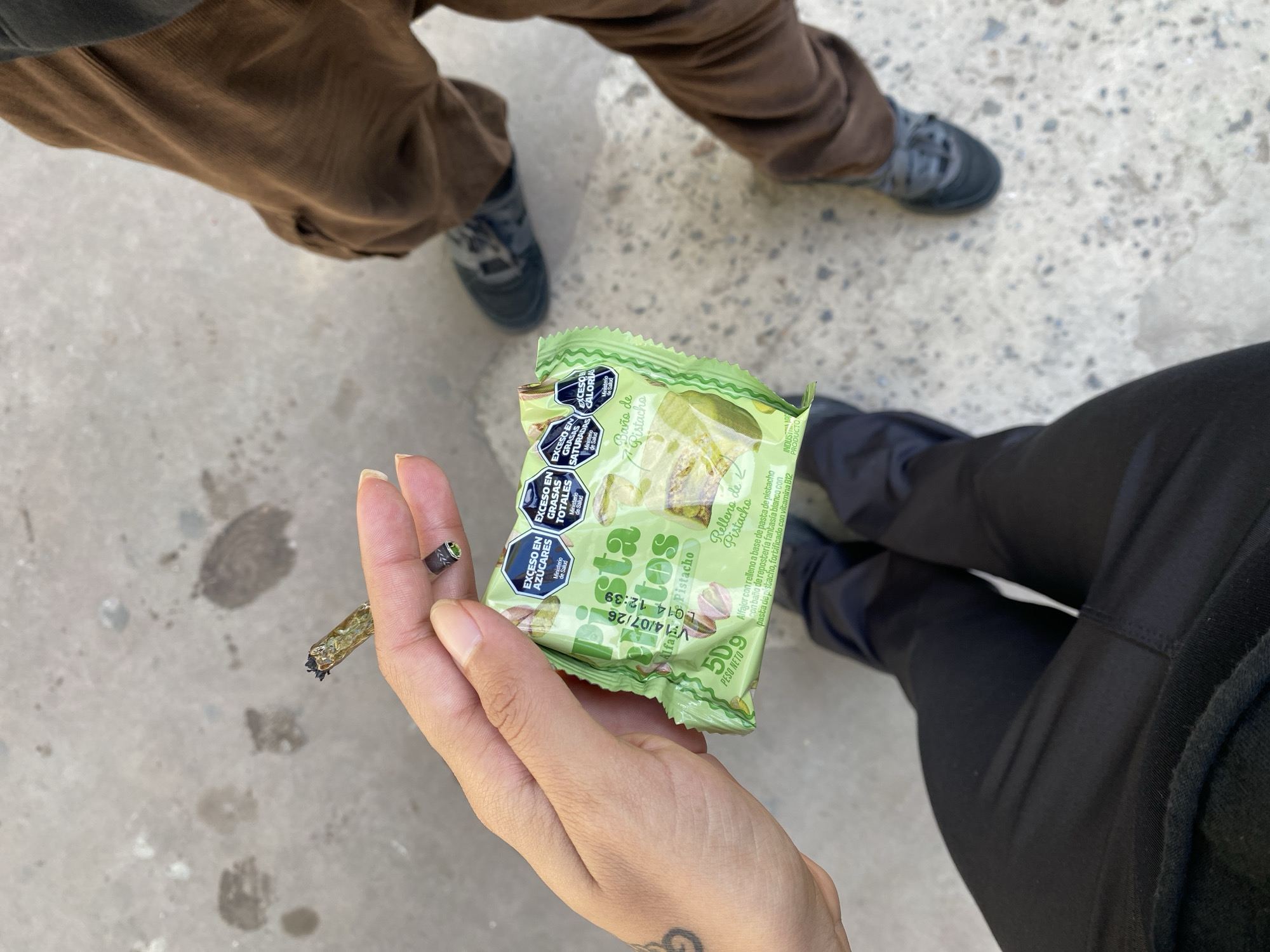 The scene presents an overhead shot of a person's hand holding a partially smoked joint, along with a green package of "Alfajor Relleno de Pistacho" candy. The packaging contains nutritional information, highlighting excess sugars, total fats, saturated fats, and calories. The setting appears to be outdoors on a concrete surface, with two other individuals standing nearby; one in brown corduroy pants and gray shoes, and another in black pants and dark shoes. There's no verifiable indication that the location is in Monte Grande, Argentina, other than the text on the candy wrapper indicating that it is an "Industria Argentina" product.
