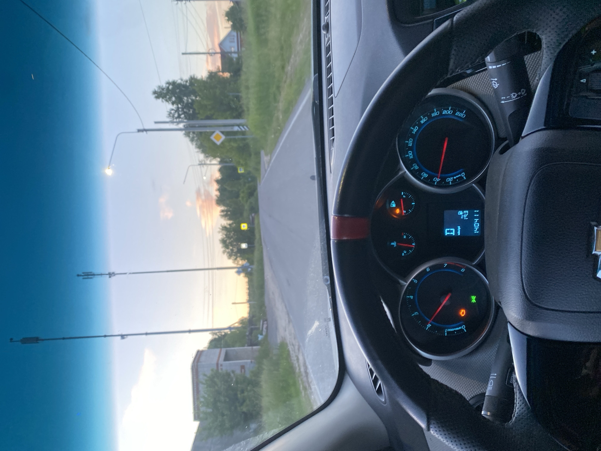 The image shows the interior of a car, looking out through the windshield. The driver's side dashboard and steering wheel are visible in the foreground. The car is on a paved road that curves to the right. In the distance, there are trees, houses, and utility poles. The sky is a light blue with some clouds, and the sun appears to be setting, casting a warm glow on the scene. The road appears to be in a rural or semi-rural area.
Based on the provided information, this scene is likely located in Медведево, Russia. The presence of a road, houses, trees, and utility poles suggests a populated area. The sky indicates it is either sunrise or sunset. The interior of the car is typical for a passenger vehicle, with the dashboard displaying the speedometer, tachometer, and other indicators. There are no people visible in the image.