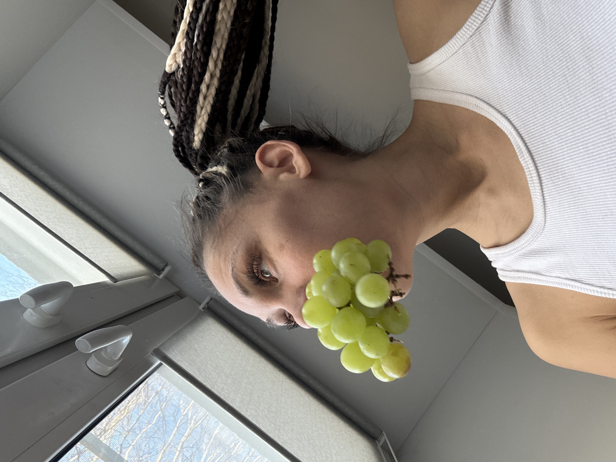 A photograph shows a person indoors holding a bunch of green grapes up to their mouth. The person has black and blonde braided hair and is wearing a white tank top. The photograph is taken from a low angle, with a window visible in the background to the left. The location appears to be indoors, but specific details about the building or location in Unknown, Unknown are not identifiable from the image.