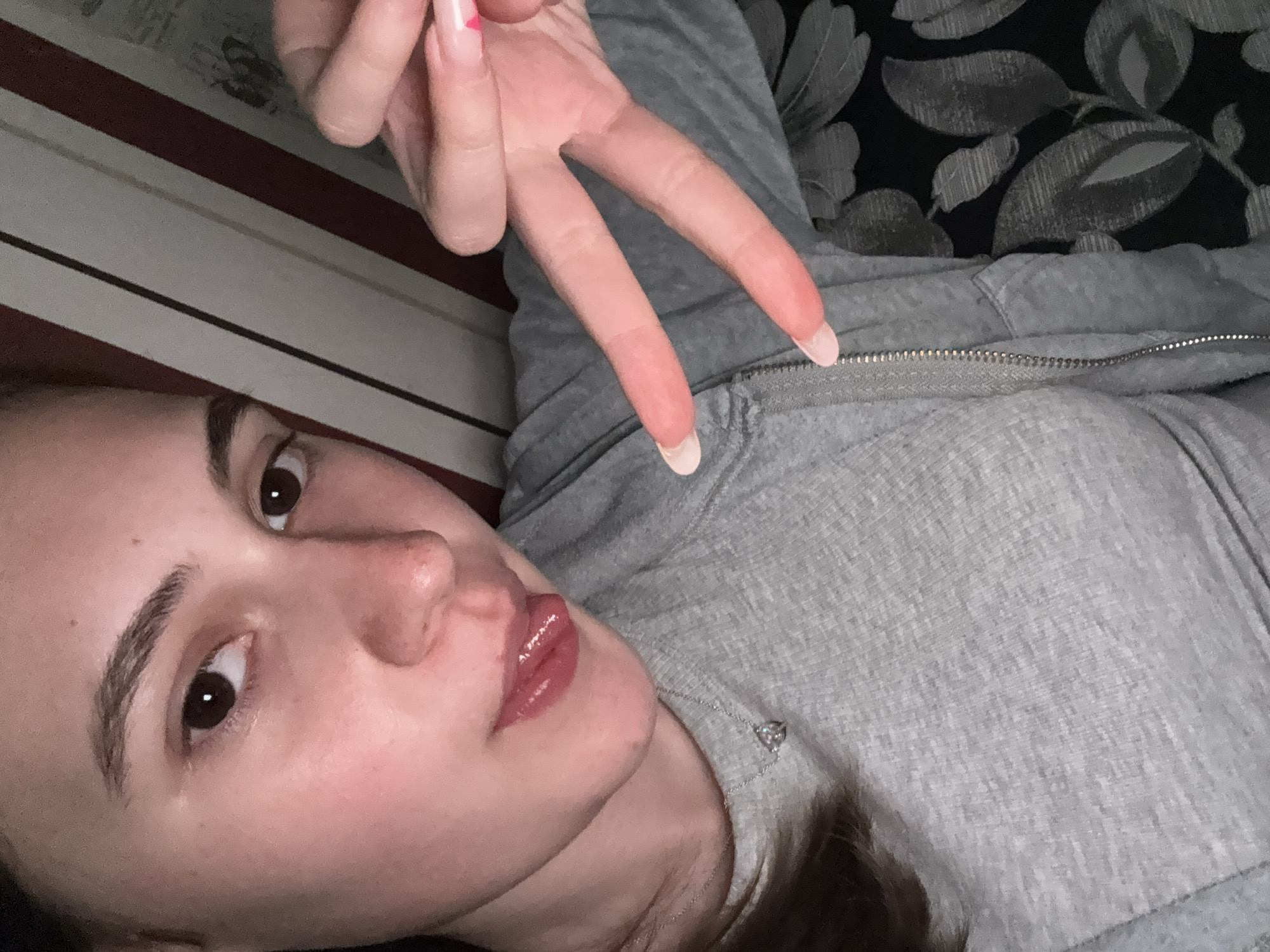 The image presents a tilted, close-up indoor portrait of a young woman, likely a selfie. She has dark brown eyes and well-defined, dark eyebrows. Her lips are full, appearing glossy with a reddish tint. She wears a grey, textured hooded sweatshirt featuring a visible silver zipper. A delicate silver chain with a small, sparkling heart-shaped pendant rests around her neck.
Her right hand is positioned in the upper center of the frame, making a "peace" or "V" sign with her index and middle fingers. Her fingernails are long and shaped into an almond or coffin style, adorned with a light pink French tip design. A distinct small red heart decal is visible on one of her upper fingernails.
The background suggests an interior setting. To the left, a surface with horizontal stripes of white and dark red or maroon is visible. To the right and behind the woman, there is a dark fabric patterned with grey floral or leaf motifs, possibly a pillow, blanket, or upholstery. The lighting is soft and even, consistent with an indoor environment. The woman's expression is calm and neutral as she looks towards the camera. No discernible text is present in the image. The context location is Panagyurishte, Bulgaria.