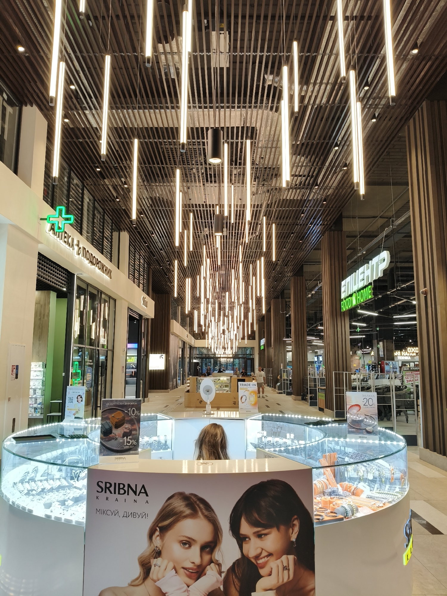 This is a medium shot of the interior of a modern shopping mall in Lviv, Ukraine. The scene is dominated by an overhead ceiling with an intricate design of parallel wooden slats and numerous vertical tube lights, creating a warm and ambient atmosphere. In the foreground, a circular jewelry display case with a glass top showcases various necklaces, bracelets, and earrings. The case is illuminated from within and around its base.
To the left of the display case is a large advertisement featuring two young women, with the brand name "SRBINA KRAINA" prominently displayed. Above the advertisement, a sign with Ukrainian text and a green cross indicates a pharmacy named "АПТЕКА "ПОДОРОЖНИК"" (Pharmacy "Podorozhnyk"). Further down the mall corridor, on the right side, a large green neon sign reads "ЕПІЦЕНТР FOOD HOME" (Epicenter Food Home), suggesting a large supermarket or home goods store.
In the background, the mall stretches out, with visible shop fronts and structural columns. A few people are present, including a woman with blonde hair leaning over the jewelry display and two figures blurred in the distance walking down the corridor. The overall impression is one of a clean, well-lit, and contemporary retail environment.