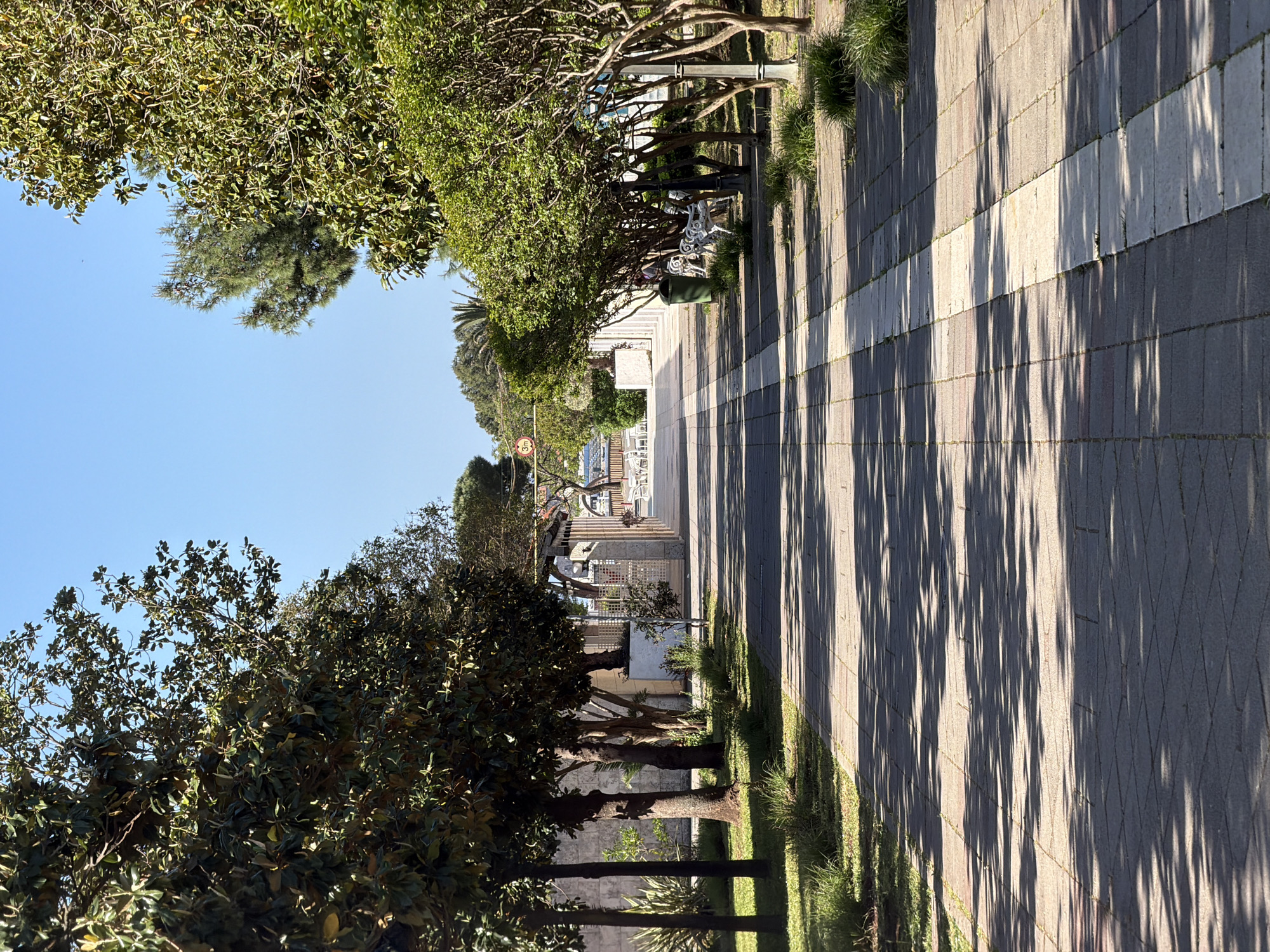 The image depicts a pedestrian walkway in Budva, Montenegro. The walkway is paved with alternating gray and white rectangular stones, creating a striped pattern. Long, dark shadows of trees stretch across the path. On either side of the walkway are trees, with dense foliage. A green trash can is visible near some white benches and white fencing. In the distance, there appears to be a building or structure with white outdoor furniture, suggesting a public space near the beach. The sky is clear and blue. A small 30 km/h sign is posted near the sidewalk.