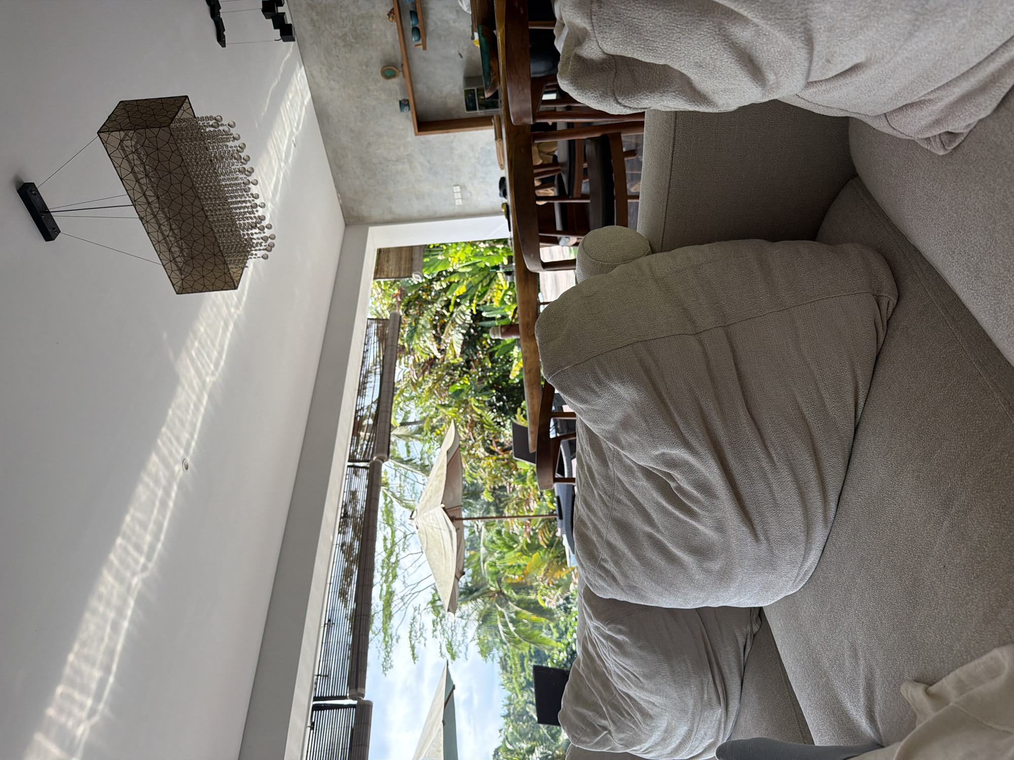The image captures an interior scene in Petulu, Indonesia, featuring a view from inside a building towards the outdoors. Prominently displayed in the foreground is a light-colored sofa with several cushions. To the left of the sofa, a wall with a chandelier hangs from the ceiling, casting light patterns. Large openings frame lush greenery and the natural environment outside. Through the doorway, there is a table and chairs that are located in a dining space.