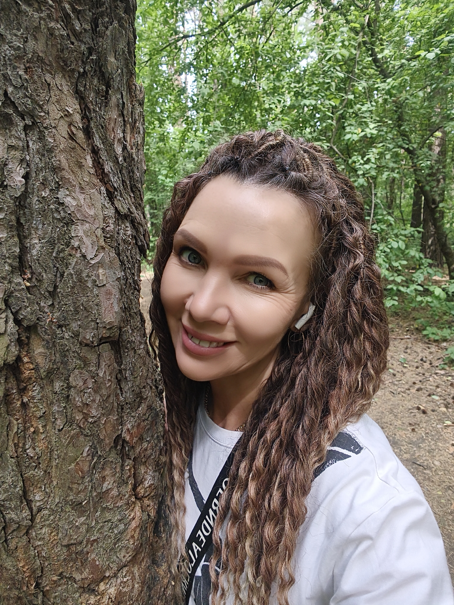 A woman with long, curly brown hair stands next to a large tree. She is wearing a white t-shirt with a black strap across her chest and a wireless earbud in her right ear. The tree has rough, brown bark. The background is a green forest with a path visible on the right. The location appears to be a wooded area.