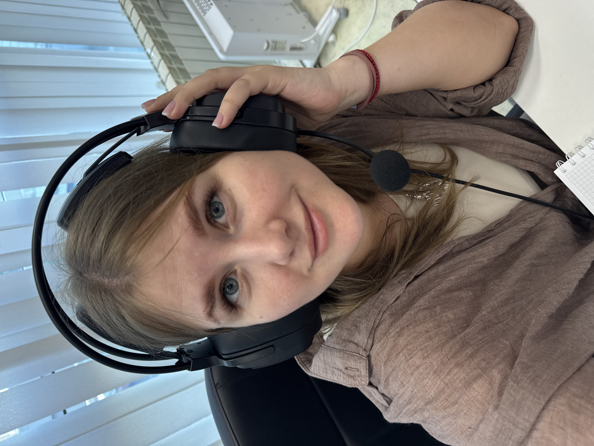 A woman is seated at a desk, wearing a black headset with a microphone. She is light-skinned with blue eyes and light brown hair. She is wearing a brown linen shirt and a thin red bracelet on her wrist. Her left hand is holding the headset to her head. A notebook with a grid pattern is visible on the desk. The background includes white blinds, a white wall, and what appears to be office equipment. The location is likely an office setting, possibly in Perm, Russia, based on contextual information, but this is not explicitly verifiable from the image alone.