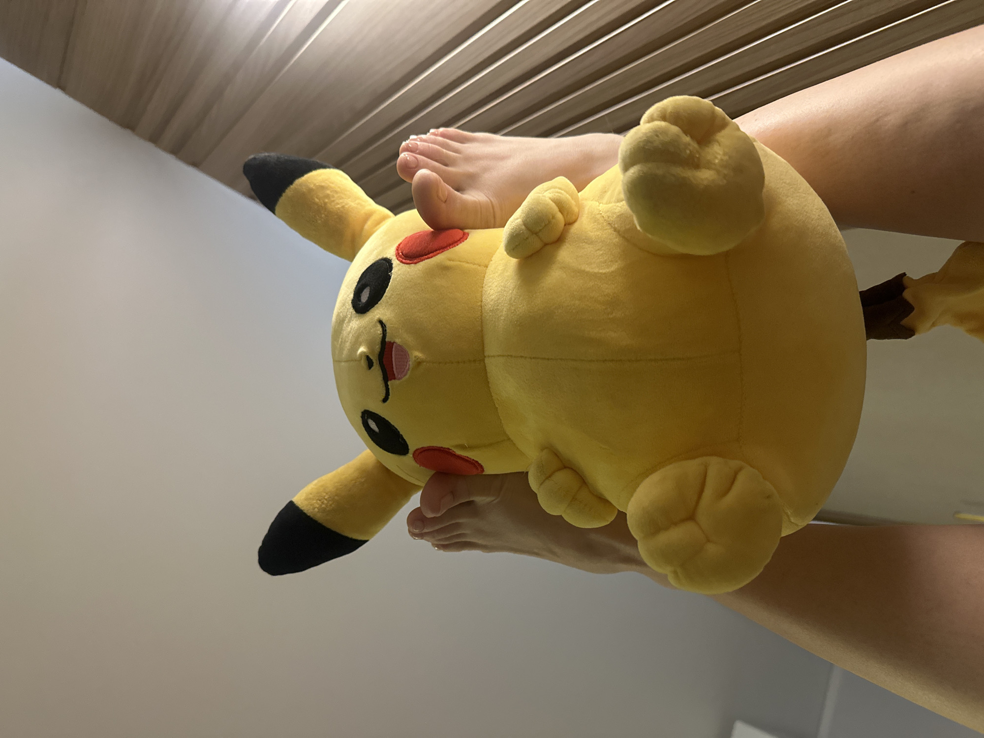 The image shows a yellow Pikachu plush toy being held in front of a person's feet. The person is lying down with their legs in the air. The background includes a wall, ceiling, and blinds. There is no information about the location being Votkinsk, Russia, or any other specific location.