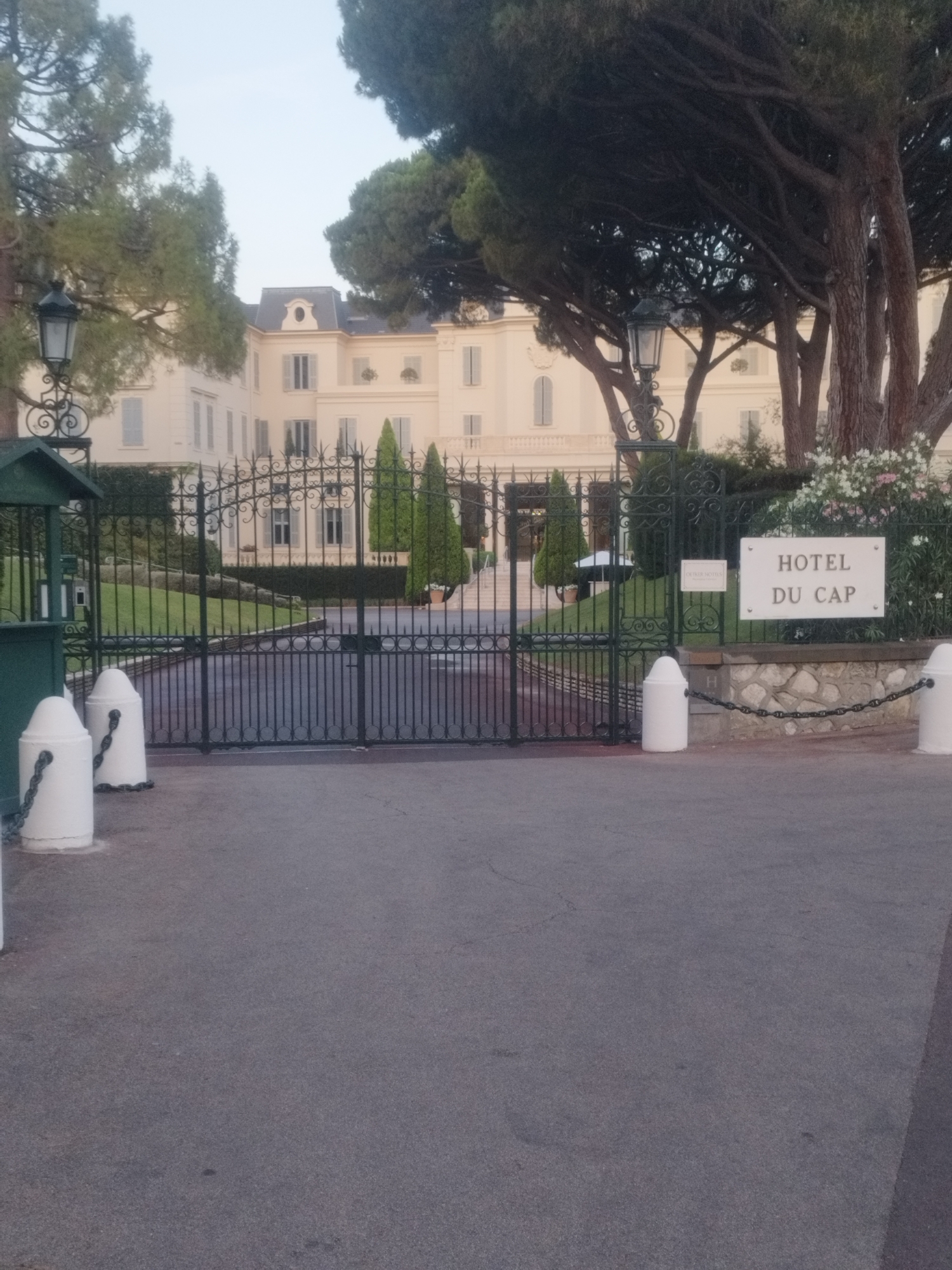 The image shows the entrance to the Hotel du Cap-Eden-Roc, a luxury hotel located on the Cap d'Antibes in Antibes, France. The main subject is the grand wrought-iron gate leading into the hotel grounds. Beyond the gate, a large, pale yellow building with a mansard roof and multiple windows is visible. The building appears to be multiple stories high and has a classical architectural style.
In front of the gate, there is an asphalt driveway. Two white bollards with chains are positioned on either side of the entrance. To the left, a small green guardhouse or kiosk is partially visible. To the right of the gate, a white sign with the words "HOTEL DU CAP" in capital letters is mounted on a stone wall, which is adorned with a chain and more white bollards.
The grounds behind the gate are landscaped with green grass, manicured hedges, and several conical evergreen trees. A large, mature pine tree with dark bark and dense needles dominates the right side of the frame, casting a shadow over parts of the scene. Flowering plants with pink and white blossoms are visible near the sign on the right. The sky is overcast and a pale blue-gray, suggesting it might be early morning or late afternoon. The overall impression is one of elegance and exclusivity.