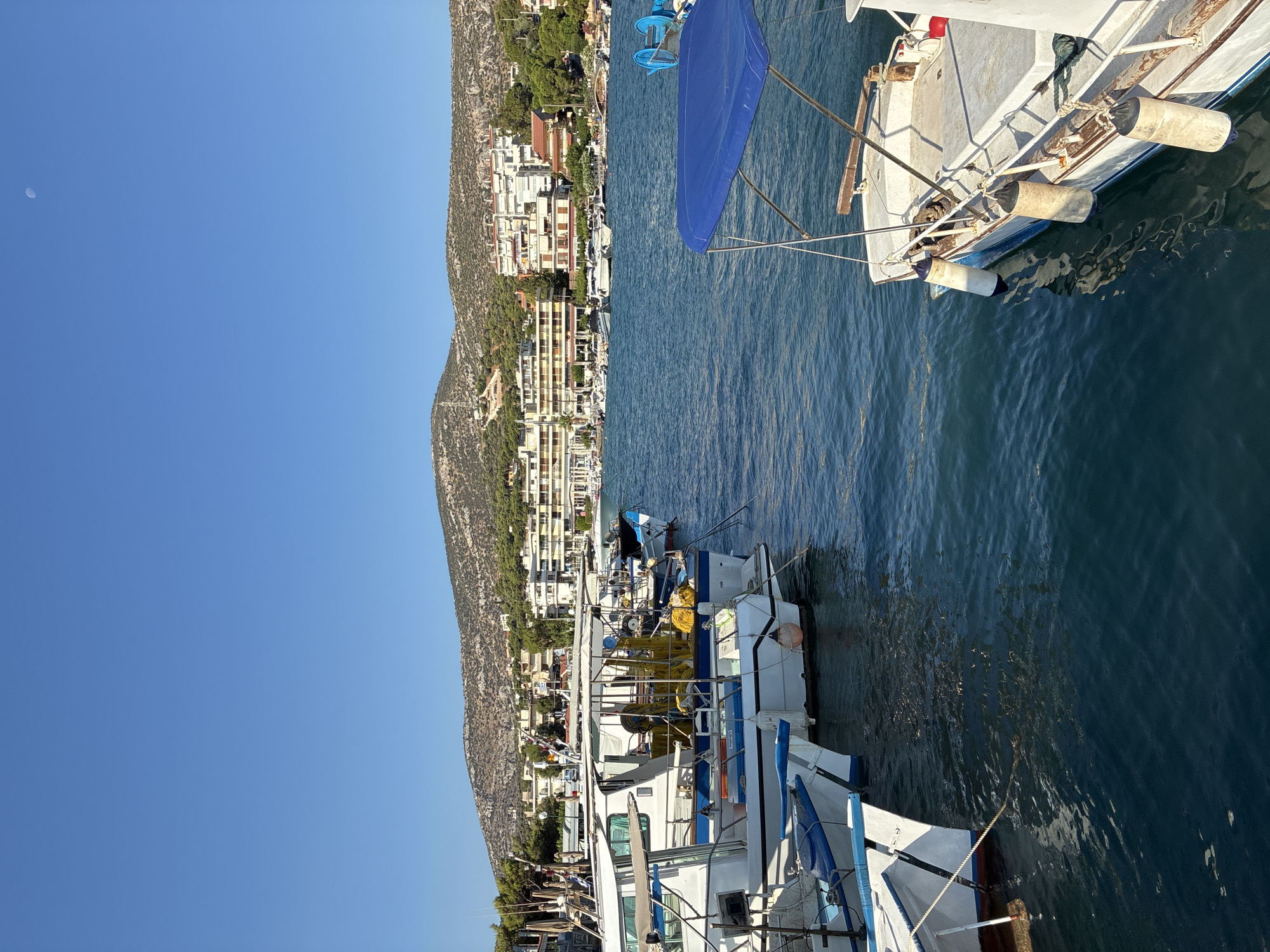 The image depicts a clear, sunny day in the harbor of Limenas Markopoulou, Greece. The foreground is dominated by several docked boats, primarily white with blue trim, some of which appear to be fishing vessels equipped with yellow netting. One boat on the right has a blue canopy. The water is a deep blue, reflecting the clear sky. In the background, a row of buildings, including hotels and houses, line the shore. Behind the buildings, a tree-covered hill rises, with the peak appearing somewhat arid and rocky. The sky is a vibrant, cloudless blue, with a faint crescent moon visible near the top left. No people are visible in the image.