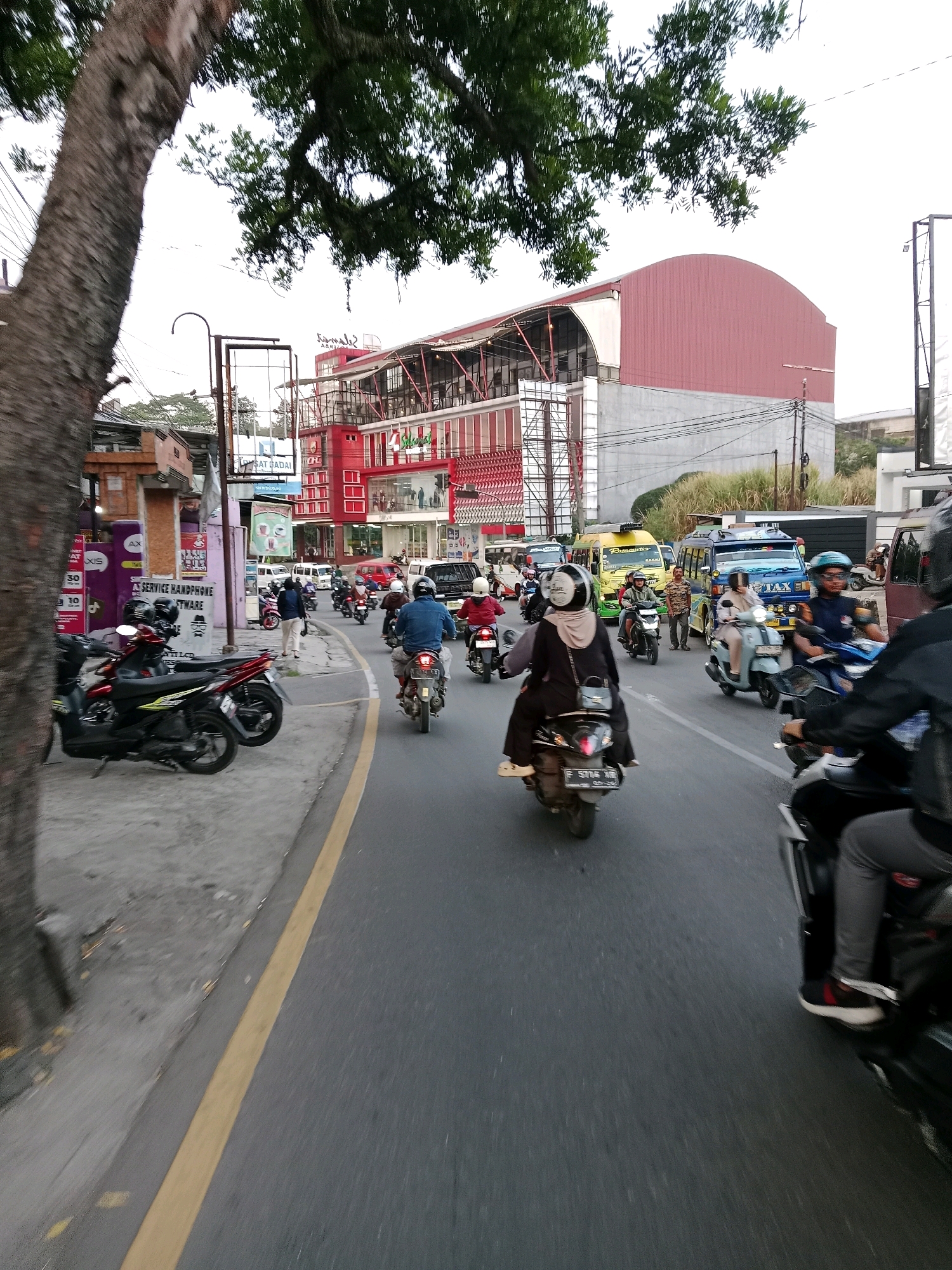 The image depicts a street scene in Sirnagalih, Indonesia, characterized by a bustling atmosphere with multiple motorbikes and a few vans. A large tree with thick branches dominates the left side of the frame, partially obscuring a red and white building complex in the background. The building appears to be a commercial establishment, possibly with multiple levels and balconies.
Several individuals are seen riding motorbikes, most wearing helmets for safety. One motorbike in the foreground carries a passenger in a dark hijab and a driver in a black jacket. Another motorbike ahead has a rider in a blue denim jacket and a helmet. To the right, a person in a black jacket and grey pants is riding a motorbike, their legs and feet visible.
Various vehicles, including a yellow van and a blue van with "TAX" written on its side, are present on the road, suggesting ongoing traffic. The road is paved and marked with a yellow line on the left side. The environment appears to be urban, with buildings lining the street and overhead power lines visible. The overall scene conveys a sense of daily life and transportation activity in an Indonesian city.