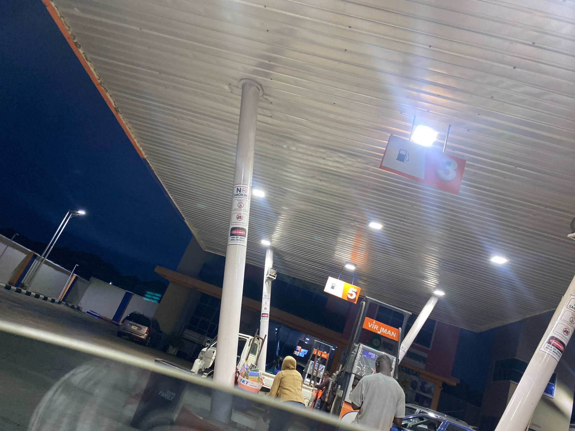 The image depicts a nighttime scene at a gas station in Jos, Nigeria. The perspective is from inside a vehicle, looking out at the gas station canopy. Multiple white pillars support the corrugated metal canopy, which is illuminated by bright fluorescent lights. Signs are visible on the pillars, including a "NO SMOKING" sign. To the right, a fuel pump displays the number "5" and the text "VIR JMAN", with digital readouts below indicating prices. Two individuals are near the fuel pump. One, wearing a light brown jacket, is facing away from the camera, while another individual, wearing a grey t-shirt, is facing the fuel pump. In the background, a white truck is parked, and to the left, a silver minivan is visible outside the covered area, along with a street lamp. The sky is dark blue, indicating it is nighttime. The overall environment suggests a typical gas station at night, with artificial lighting highlighting the operational areas.
