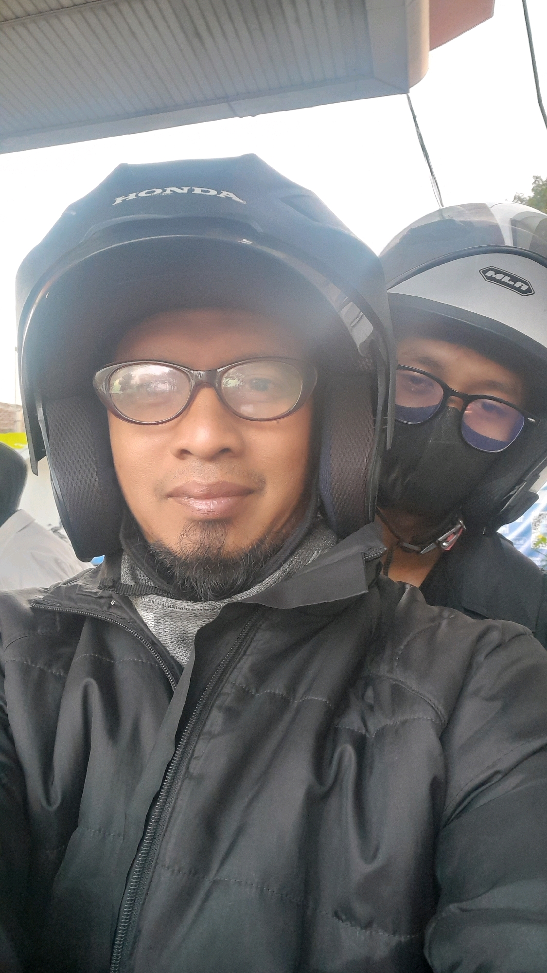 Two individuals wearing helmets are seated on a motorcycle, likely for transportation. The person in the foreground is wearing glasses and a black jacket, with a goatee. The second individual, partially visible behind the first, is also wearing a helmet and glasses, and a dark face mask. The background appears to be an outdoor setting with a covered structure overhead.
