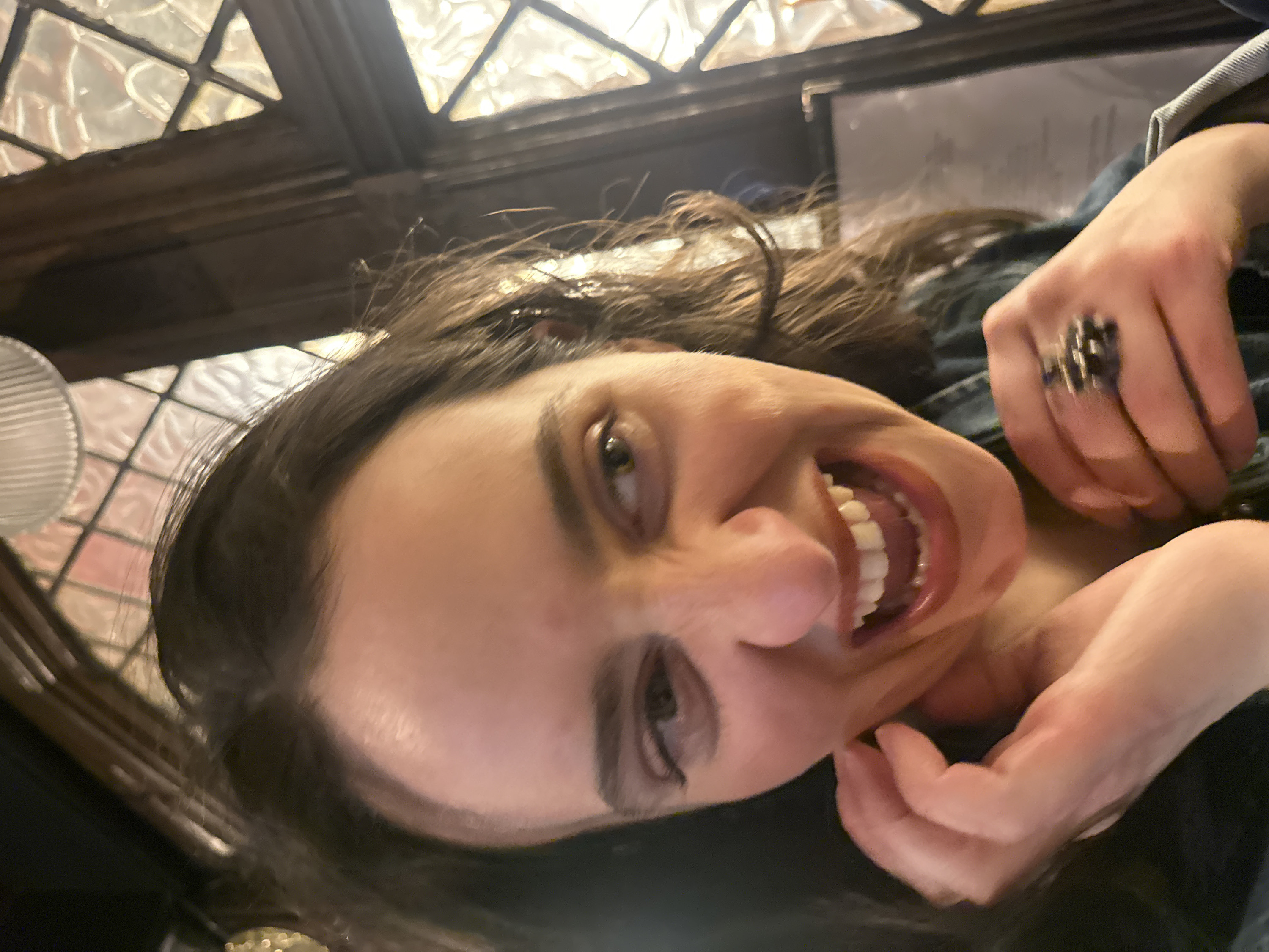 A woman with long brown hair smiles widely, her head tilted. Her left hand is near her chin and the right hand is above her. A large, ornate ring adorns the middle finger of her right hand. She wears a blue denim jacket. Stained glass windows with a dark frame are visible in the background, with a white, possibly translucent, object in front of them. The lighting is somewhat dim, creating shadows across her face.