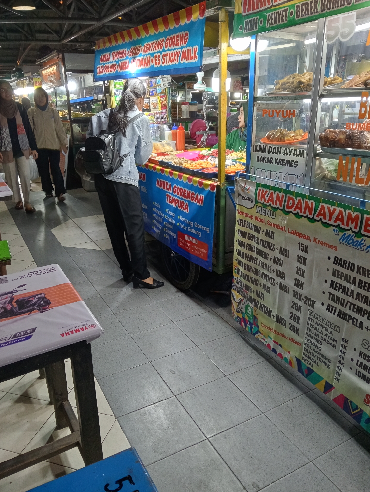 The image captures a street food stall in Magelang, Indonesia, specifically identified by the "Nageland" text on the menu banner. The stall features a variety of fried foods and grilled chicken and quail, as indicated by the signage. In the foreground, a young woman with grey hair is standing, facing away from the camera, and appears to be a customer. She is wearing a denim jacket, black pants, and black heels. Behind her, two other women, dressed in casual attire, are also visible, one in a hijab and the other in a beige jacket. The stall is illuminated by overhead lighting, and the floor is tiled with grey and white squares. The menu board displays a list of food items and their prices, written in Indonesian. The overall scene depicts a bustling food market or street food area.