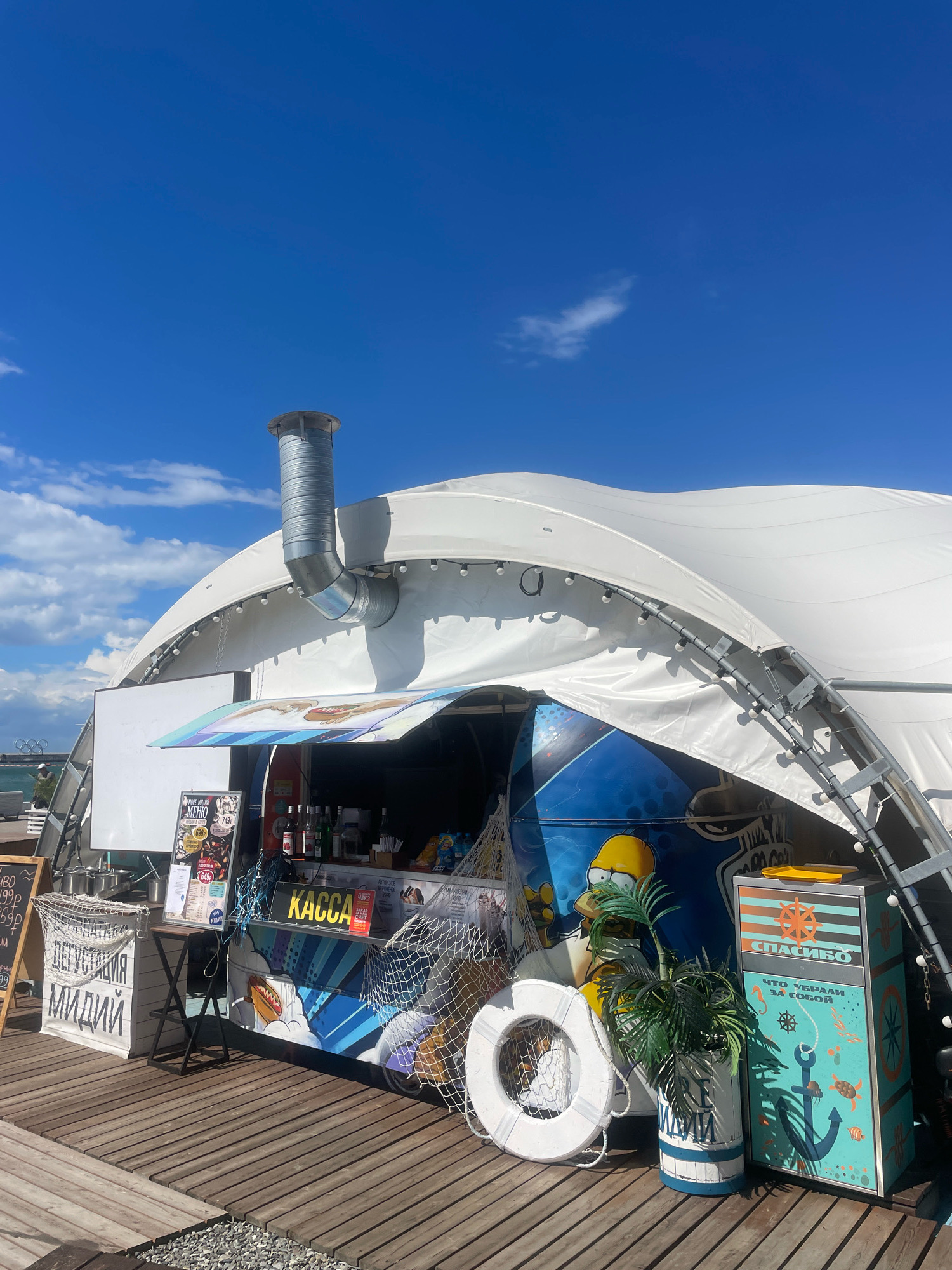An outdoor food kiosk, decorated with images of Homer Simpson, a hot dog, and nautical elements, is situated on a wooden boardwalk under a large, white, arched tent. The kiosk has a counter with various bottles and containers, a "KASSA" (cashier) sign in yellow, and a menu board to the left. To the right of the kiosk, there is a colorful trash receptacle with the word "СПАСИБО" (thank you) and a drawing of an anchor, lifebuoy, and marine life. A potted palm tree stands next to the trash can. In the background, the sea and a clear blue sky with some clouds are visible. The scene appears to be in a public space, likely a promenade or beach area, given the boardwalk and proximity to the water. The overall atmosphere is bright and summery. There is a man in the background on the left side of the image. The kiosk is likely a vendor selling food and drinks.