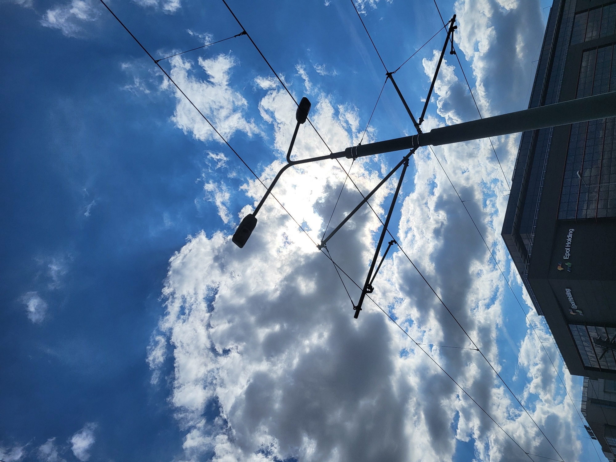 The image, taken from a low angle, shows a view towards a cloudy blue sky in Unknown, Unknown. Dominating the foreground is a metal pole and overhead power lines, typical for tram or train electrification. Attached to the pole are two streetlights. To the right of the frame, the corner of a building is visible with the inscription "Epol Holding". The sun is partially obscured by clouds. The absence of people and cars indicates a possible moment of pause or a setting outside peak activity periods. The image suggests an urban environment with tram or train infrastructure.
