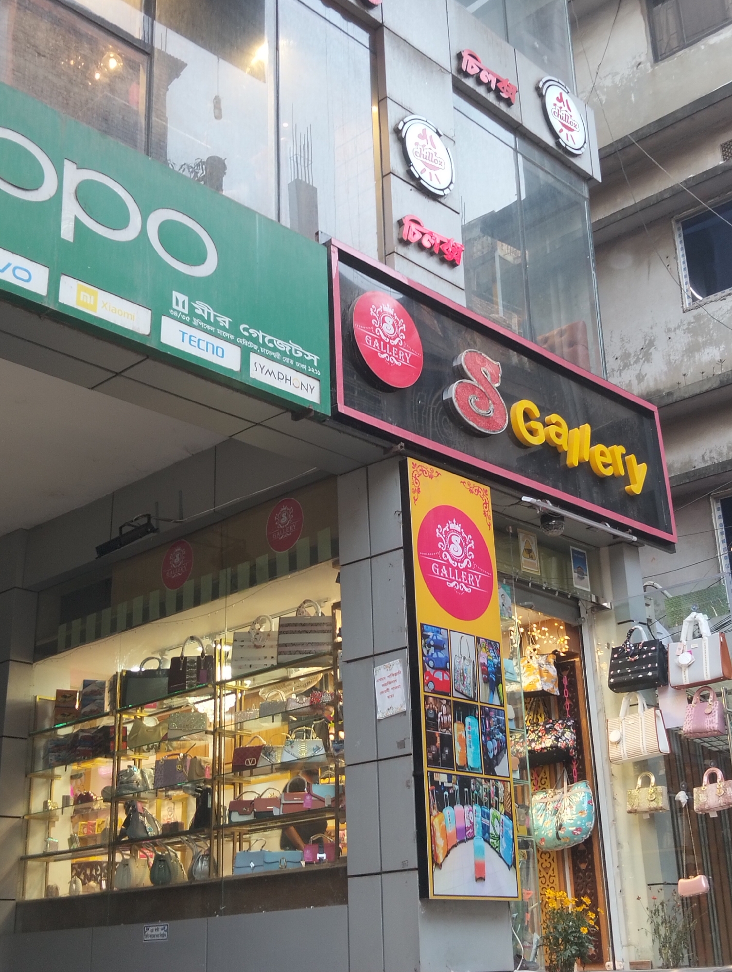 The image captures the exterior of a commercial building in Dhaka, Bangladesh. Several businesses are visible. An OPPO advertisement overhangs "S Gallery," a handbag shop showcasing a variety of bags in its storefront window and on display outside. Above OPPO are signs advertising "Chillox" in both English and Bengali. The location is identified by text on the OPPO advertisement indicating that the shop is located at 34/35 Tropical Malek Heritage on Dhakeshwari Road in Dhaka 1211. Visible beyond the store is an adjacent building with a weathered exterior.
