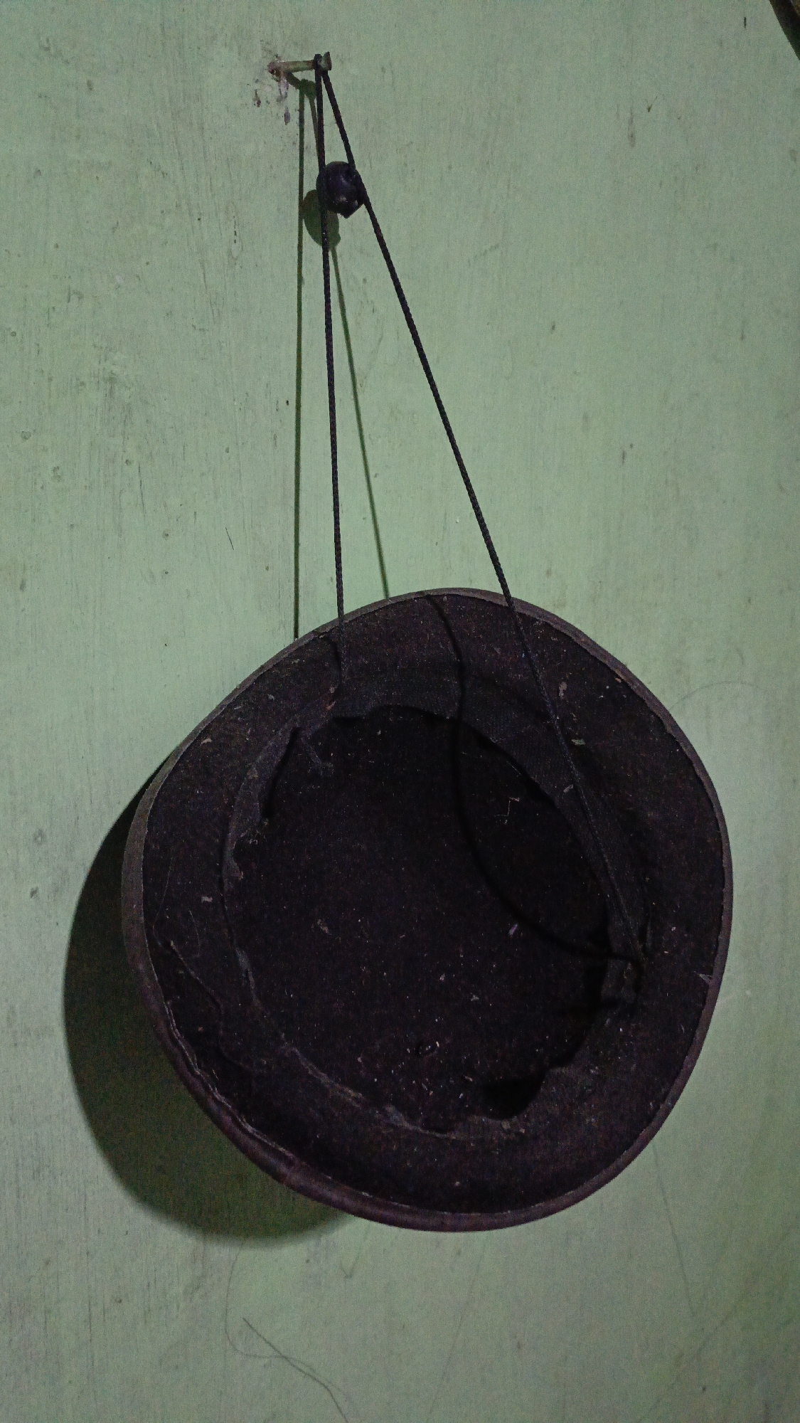 A dark-colored hat is hanging from a nail on a light green wall in Trihanggo, Indonesia. The hat is suspended by a black string that is attached to a small, dark object, possibly a bead or a stopper, which is then threaded through a loop on the hat. The hat appears to be a fedora or a similar style with a brim. The wall has a textured and slightly distressed appearance. A shadow of the hat is cast on the wall to its left. The lighting suggests an indoor environment, possibly a room.