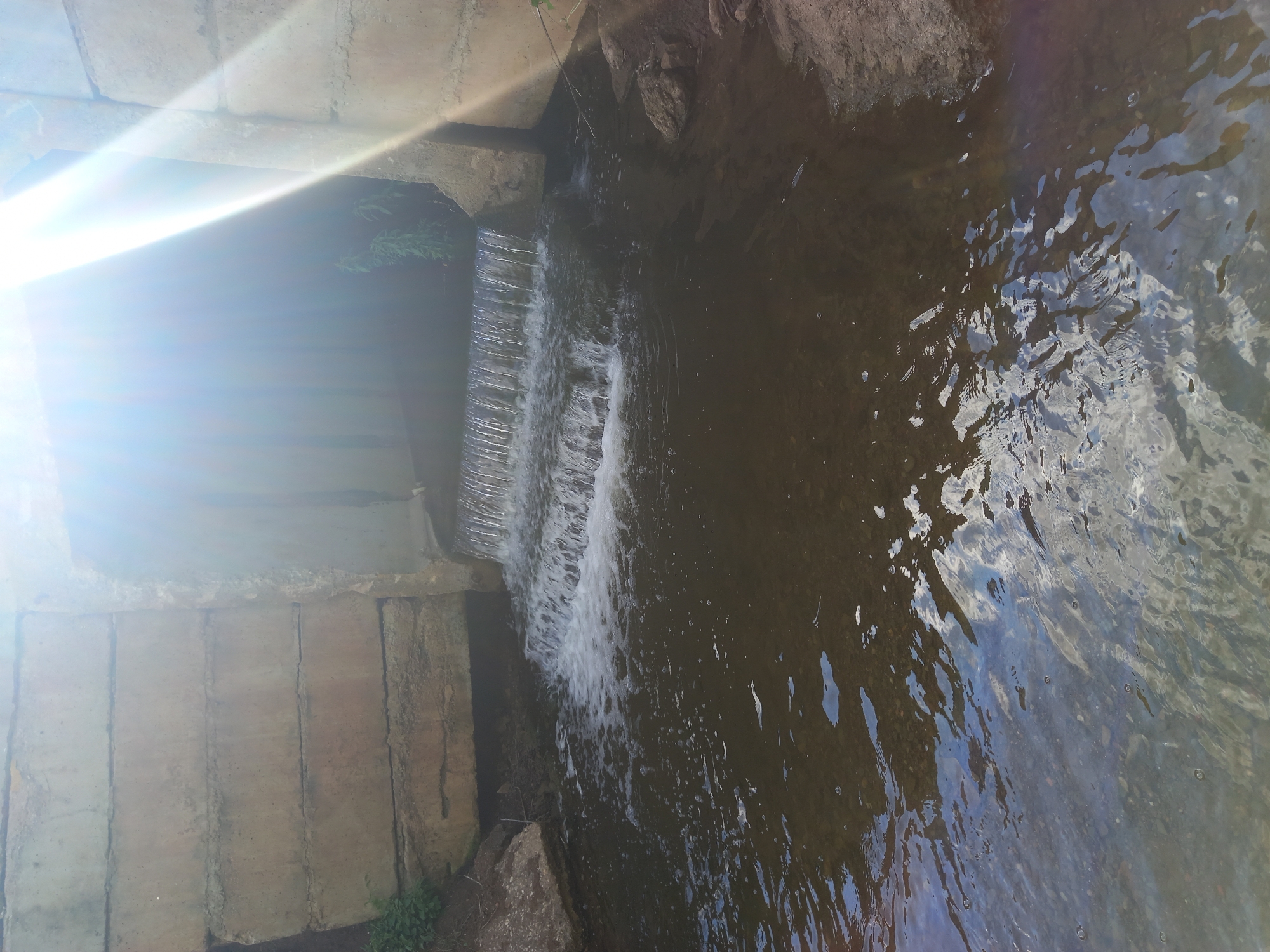 The image shows a small waterfall in a narrow channel, likely a stream or culvert, surrounded by concrete and natural rock. The water is flowing over a series of small drops, creating a white foamy cascade. The water itself is dark and appears to have a rocky or muddy bottom visible in some areas. The reflections on the surface of the water suggest a bright, possibly cloudy sky.
On the left side of the frame, there are concrete structures that appear to be part of an embankment or bridge. The concrete is weathered and shows signs of age. A strong sun glare is present in the upper left quadrant of the image, obscuring some details and casting a bright light across the concrete. Hints of green foliage are visible in the background, suggesting a natural or semi-natural outdoor setting.
The perspective is from above and looking down into the channel. The overall scene is one of flowing water in a man-made and natural environment. The time of day appears to be daytime, given the strong sunlight and reflections. There is no text visible in the image. The specific location is Sterlibashevo, Russia.