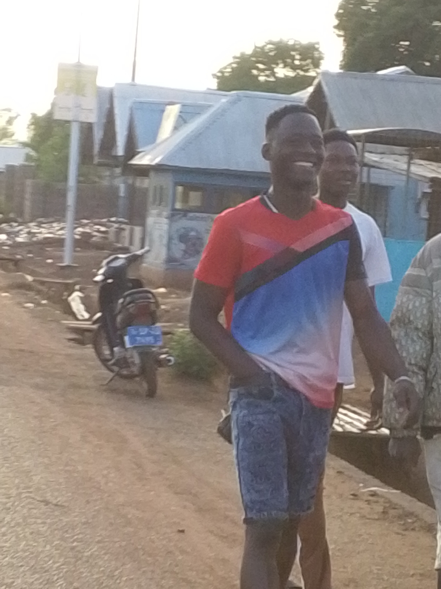 The image depicts three individuals on a dirt road, with a primary focus on the first figure who is smiling. He is wearing a t-shirt with red, black, and blue sections, coupled with patterned denim shorts. Behind him stands a second individual in a white t-shirt, partially visible. The third person is only partially in view, to the right, and is wearing a patterned jacket. A parked motorcycle can be seen in the background, near a signpost. Buildings with blue roofs and a water tank are in the background, indicative of a residential neighborhood setting. The setting suggests a location in the outskirts of Tamale, Ghana.