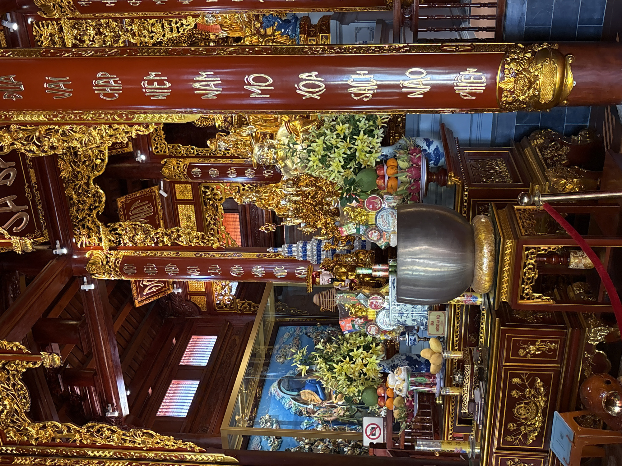 The image shows the interior of a temple in Nam Nha Trang Ward, Vietnam. The architecture is ornate with extensive use of dark wood and gold embellishments. A large wooden pillar with gold lettering dominates the foreground. Behind it, an altar displays various offerings including flowers, fruit, and religious objects. The scene is richly detailed with carvings and decorative elements, suggesting a place of reverence and tradition.