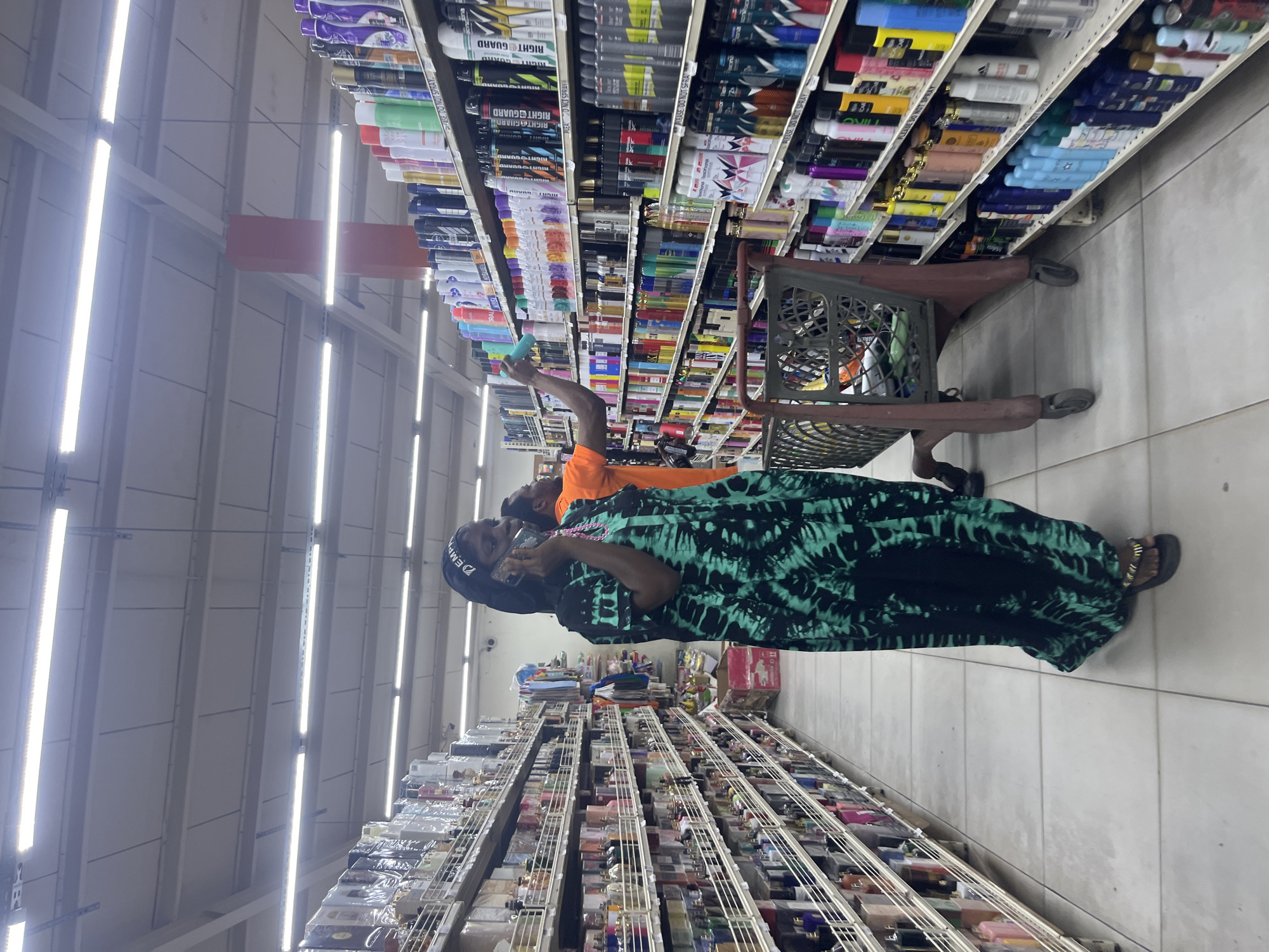 In Ado, Nigeria, two individuals are observed in a brightly lit supermarket aisle, with shelves stacked high with various products. The aisle is made of light-colored tile. A woman on the left, wearing a black and green tie-dye dress and a head covering, is speaking on her cell phone. To her right, a man in an orange shirt is reaching for a product on a shelf. A shopping cart with a red frame is positioned between them. The shelves display a wide assortment of bottled goods, including what appear to be personal care items such as deodorants and lotions. The lighting suggests it is daytime, with long fluorescent tubes illuminating the space.