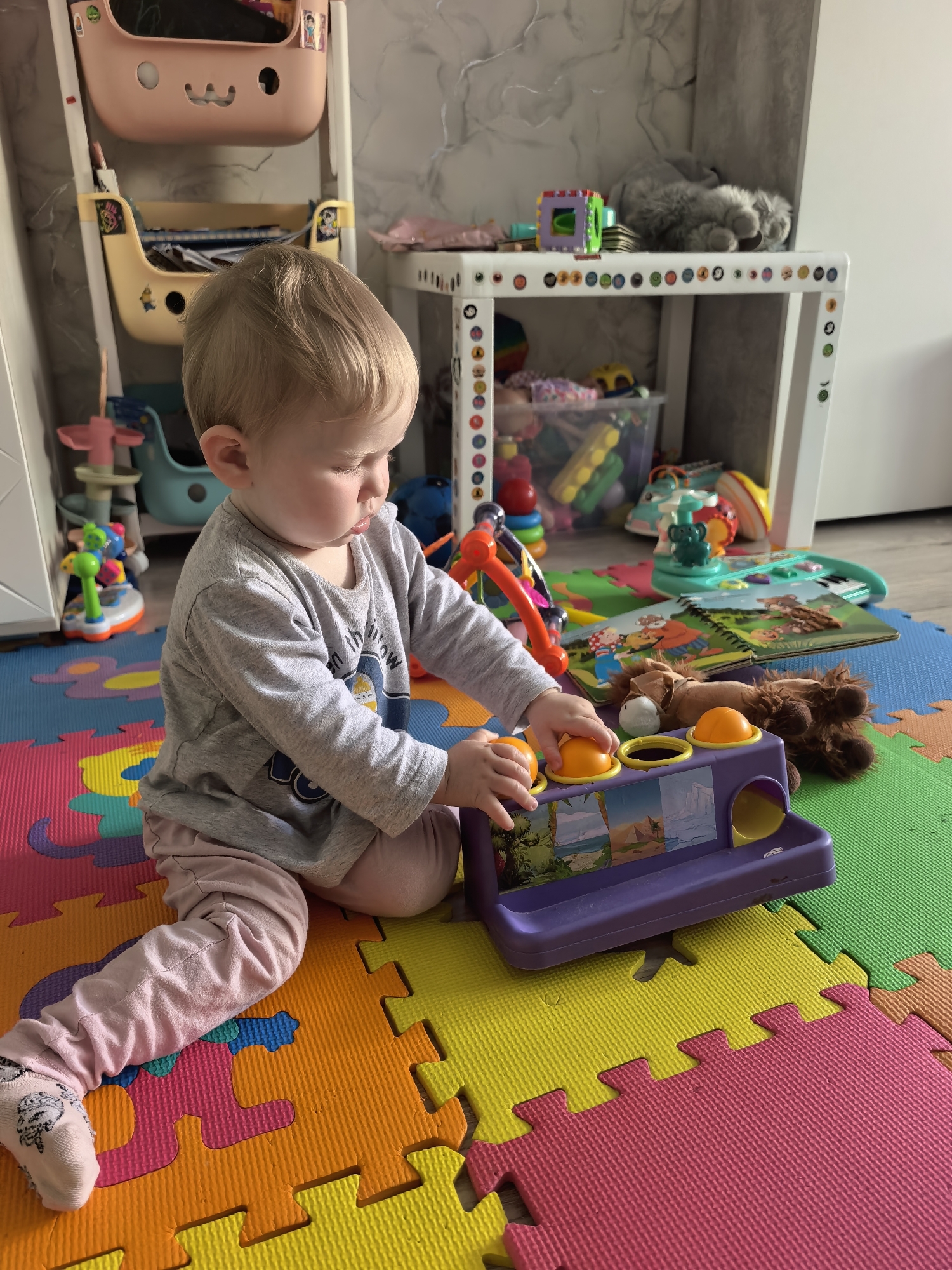 A young child with blonde hair and light skin is sitting on a colorful, interlocking foam puzzle mat and playing with a purple toy. The child is wearing a grey long-sleeved shirt with a graphic design on it and pink leggings. The toy has several yellow balls and a series of colorful pictures displayed on it. Various other toys are scattered around the child, including a stuffed animal, a book, and other plastic toys. In the background, there is a white shelf unit filled with more toys and a stuffed grey cat lying on top. The scene appears to be indoors, likely in a playroom or child's bedroom, with a textured wall and light coming from the left side of the frame, suggesting it could be daytime. The child appears focused on the toy, and there are no clear indications of specific weather conditions or emotional states beyond concentration. There is some visible text on the child's shirt, but it is not clearly legible.
