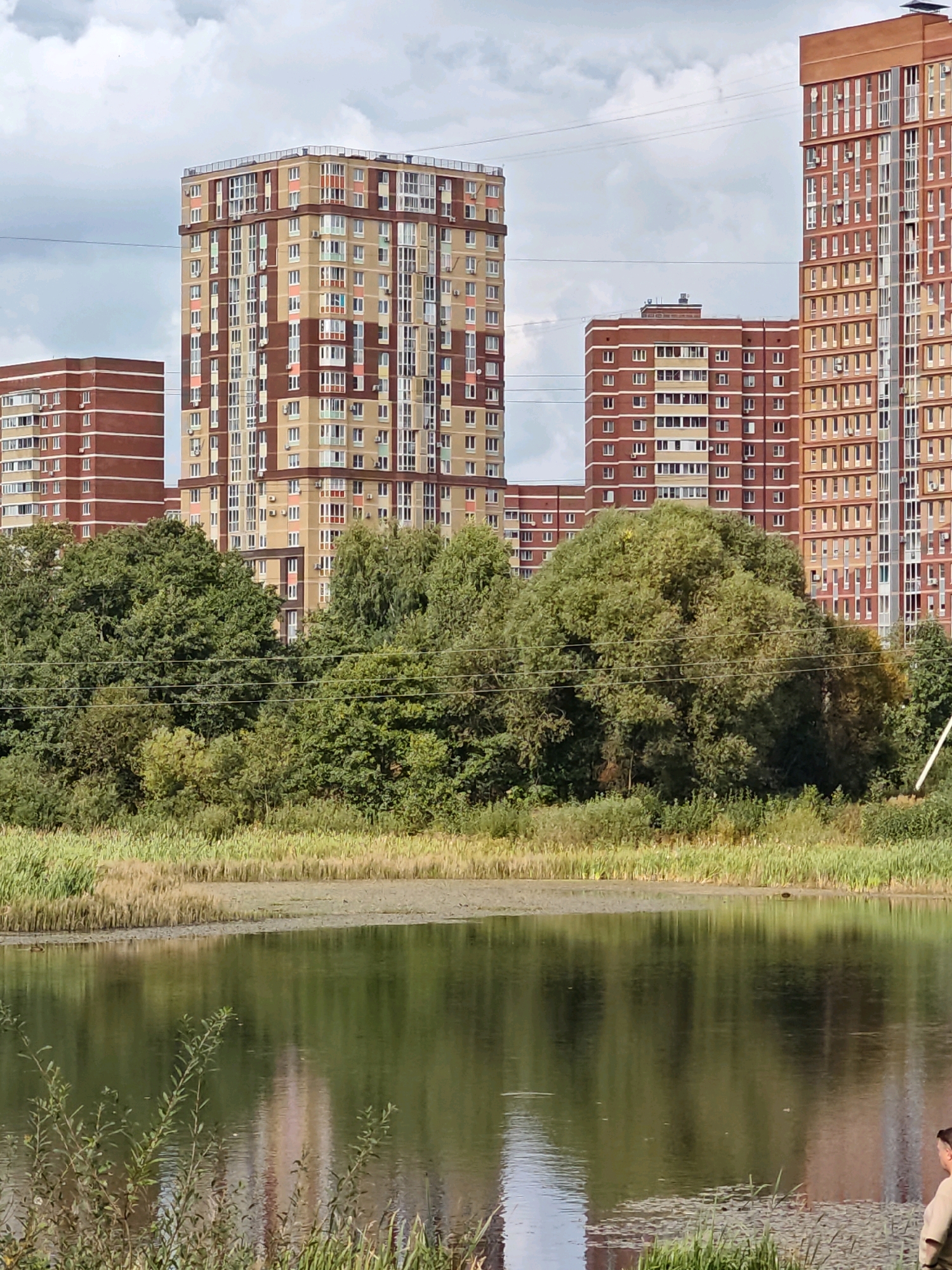 The image depicts a body of water reflecting tall, modern apartment buildings under a cloudy sky. The buildings are predominantly red and beige, with numerous windows and balconies. Green trees and foliage line the shore of the water, creating a natural buffer between the buildings and the water. The scene appears to be set in a residential area with a mix of urban development and natural elements. A person is visible in the lower right corner of the image, looking towards the water. This is an urban landscape in Shcherbinka, Russia, characterized by high-rise residential architecture and a nearby natural body of water.