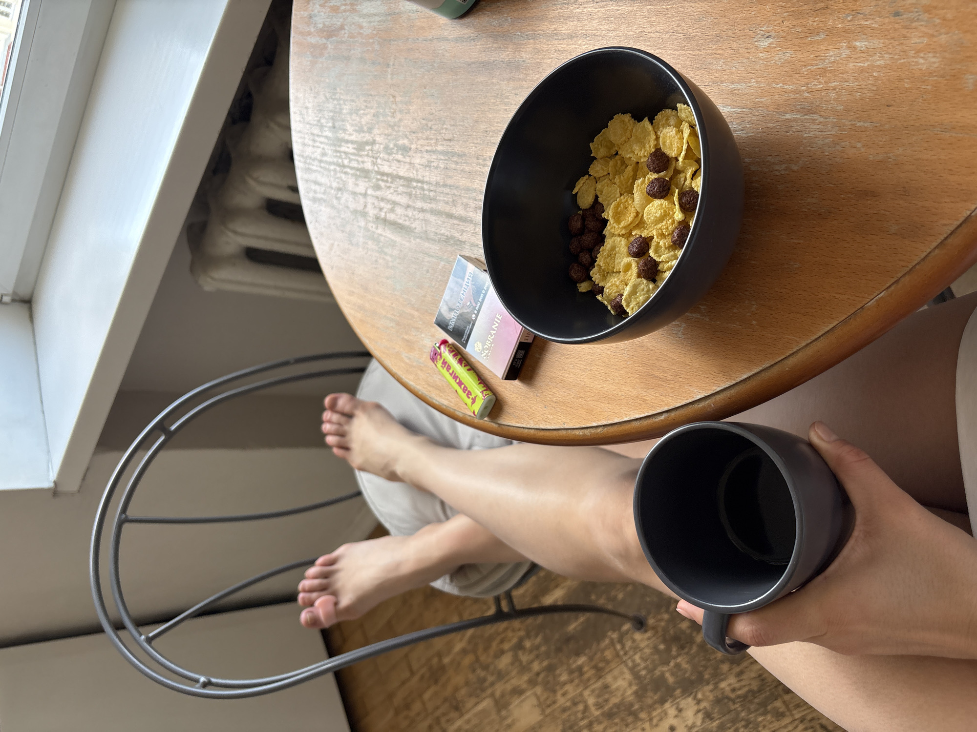 A person sits at a round, wooden table near a window and a white radiator. The person has their legs crossed and is holding a gray mug in their right hand. On the table is a black bowl filled with cornflakes and chocolate cereal pieces, a package of "Sobranie" cigarettes, and a green "зажигалка" (lighter). The scene appears to be indoors, possibly in an apartment. There's no definitive information to pinpoint the location within Moscow.