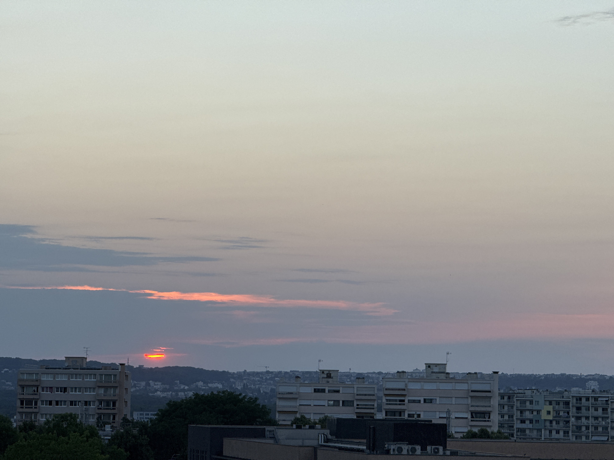 The image shows a cityscape at dusk. The sky is a gradient of pale yellow and light blue, with streaks of pink and orange clouds. A thin, bright orange horizontal line, likely the setting sun, is visible near the horizon. In the foreground, there are several multi-story buildings, some appearing to be apartment complexes with numerous windows. Trees are visible in the lower portion of the frame, and a distant, hilly landscape can be seen behind the buildings. The overall impression is that of a residential area at the end of the day.